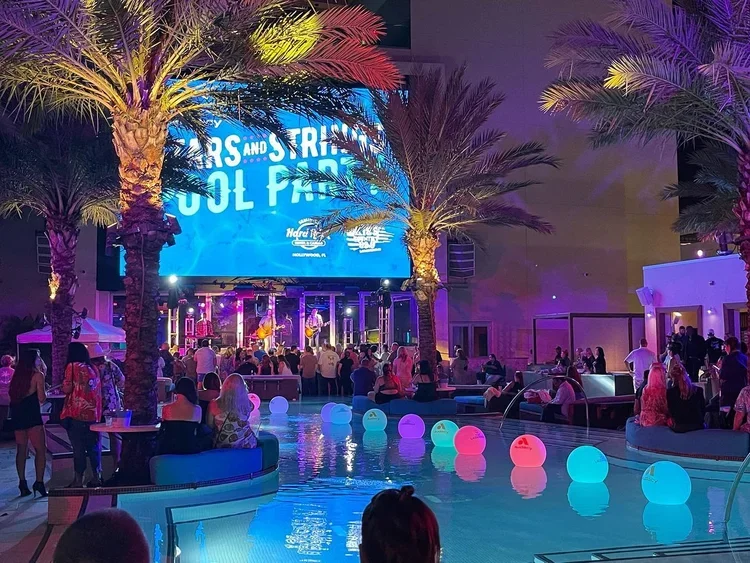 Outdoor poolside party with people, colorful lighting, palm trees, and a large screen displaying text