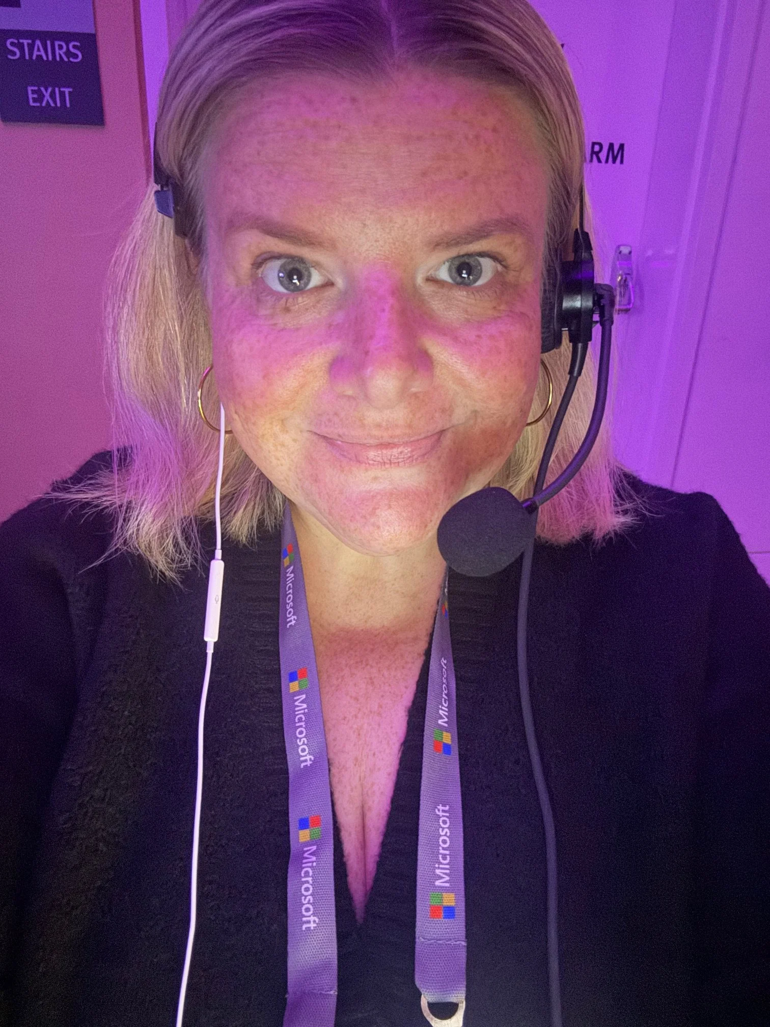 Selfie of a woman with blonde hair wearing a headset with a microphone, a black top, and a Microsoft lanyard, taken in a setting with purple lighting.