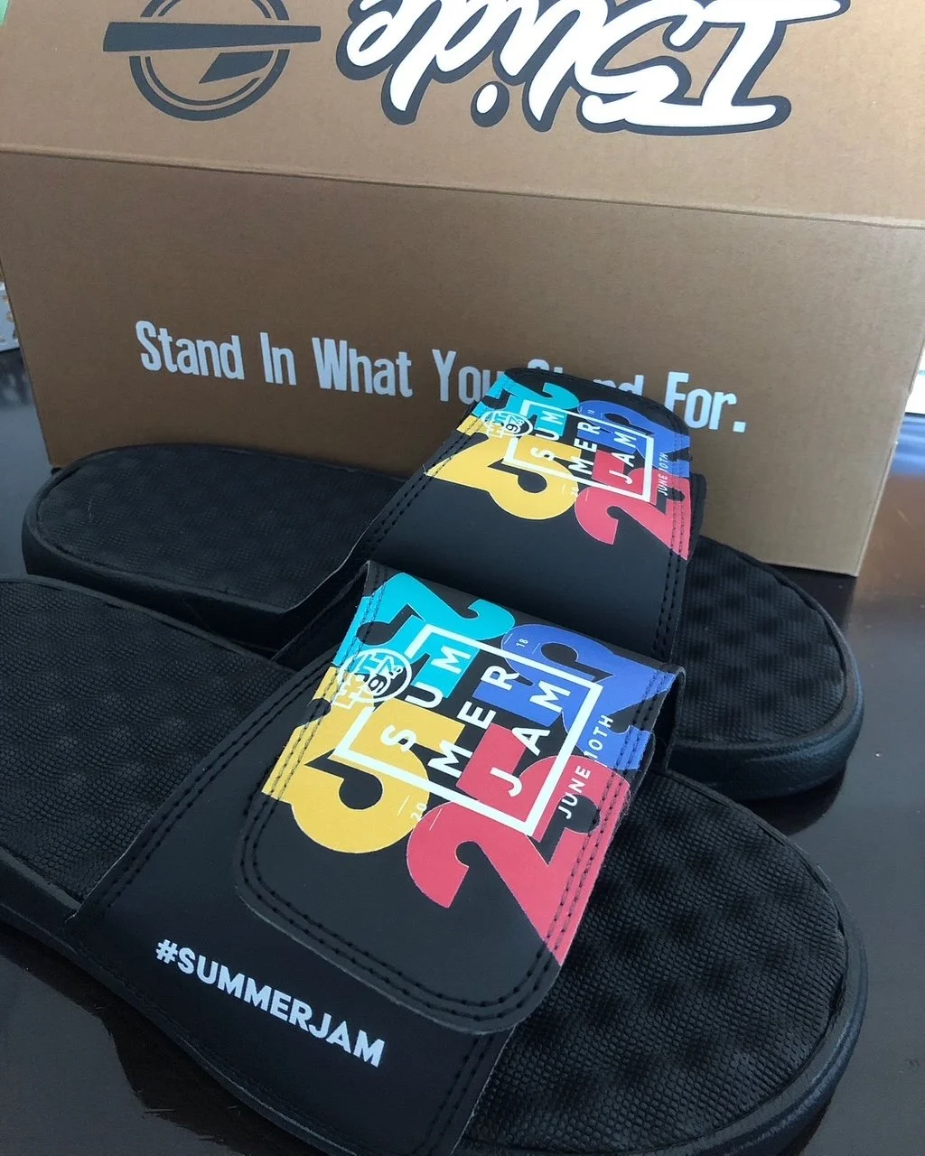 Black slides with colorful logos and the hashtag #SUMMERJAM in white, placed in front of a cardboard box with a logo and the words 'Stand in what you stand for'.