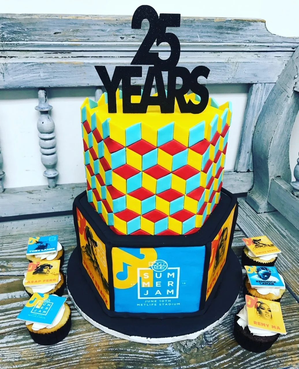 Colorful birthday cake with a yellow, red, and blue geometric pattern, topped with a "25 Years" cake topper, surrounded by cupcakes decorated with images of popular rappers and musicians, celebrating a 25th anniversary or birthday.
