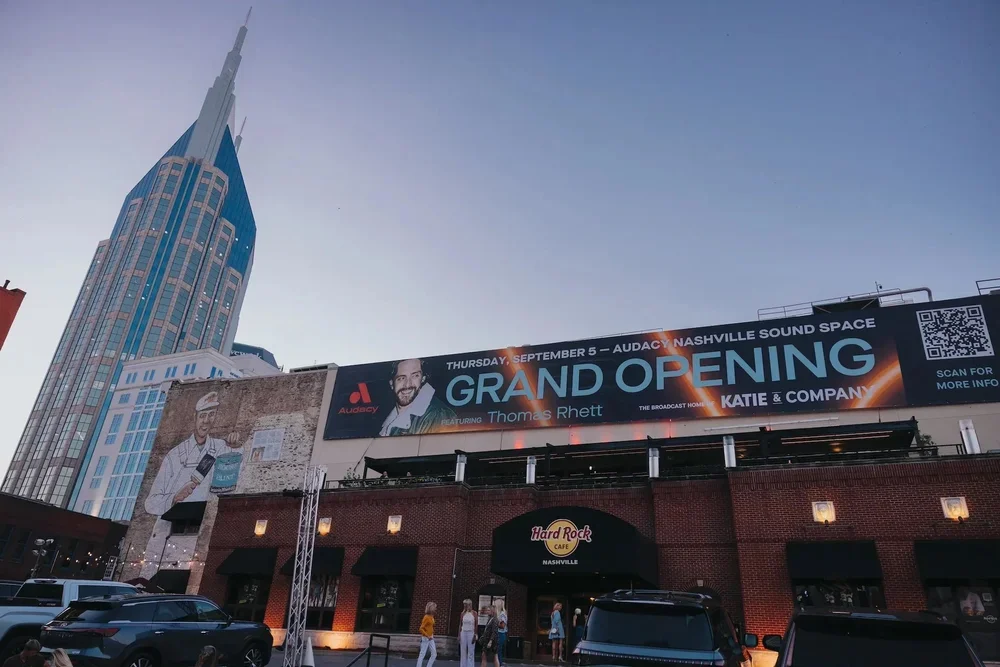 Nashville city street with the AT&T building, a large billboard announcing the grand opening of Audacy Nashville Sound Space on September 5 featuring Thomas Rhett and Katie & Company, and people walking near the Hard Rock Cafe.