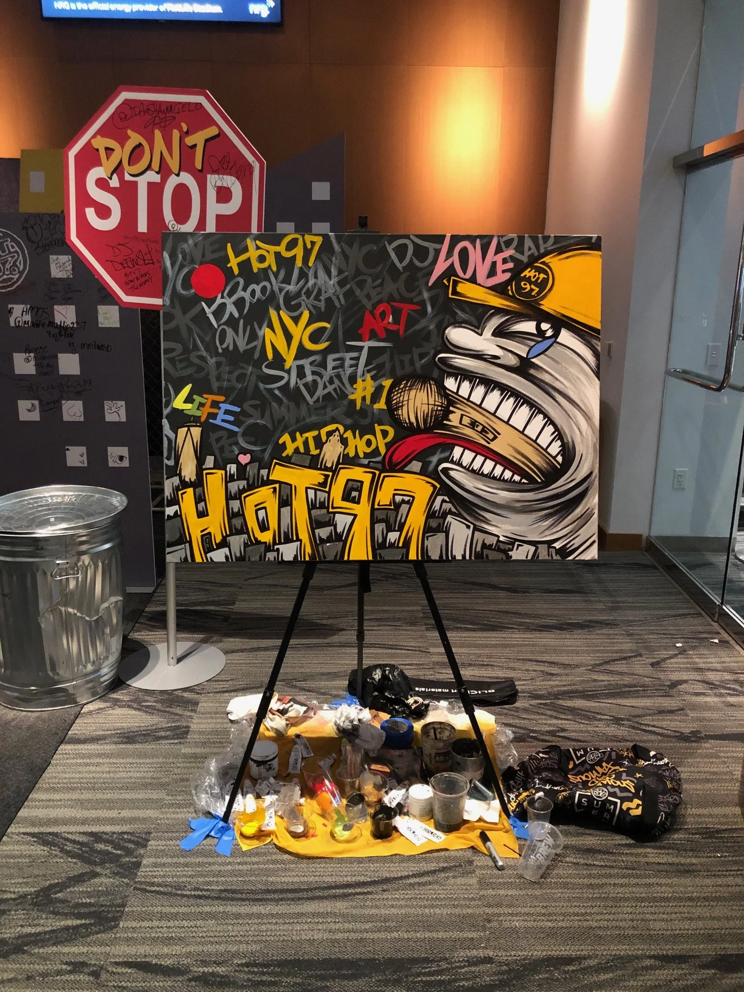 Artwork in progress with spray paint cans, paint spilling on the floor, graffiti-style design featuring a large fierce face, urban landscape, and words like 'Love,' 'NYC,' 'Hip Hop,' and 'Hot 97,' set up on an easel in a gallery or studio space.