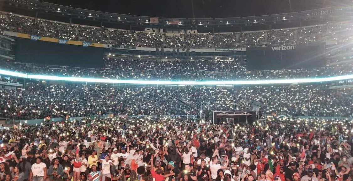 Crowd at a concert or event in a large stadium, holding up phones with flashlights on, creating a sea of lights.