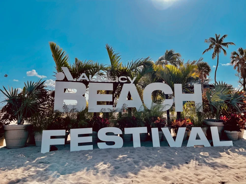 Large white sign reading "Augustine Beach Festival" set on sandy beach with palm trees and tropical plants in the background, under a blue sky with a few clouds.