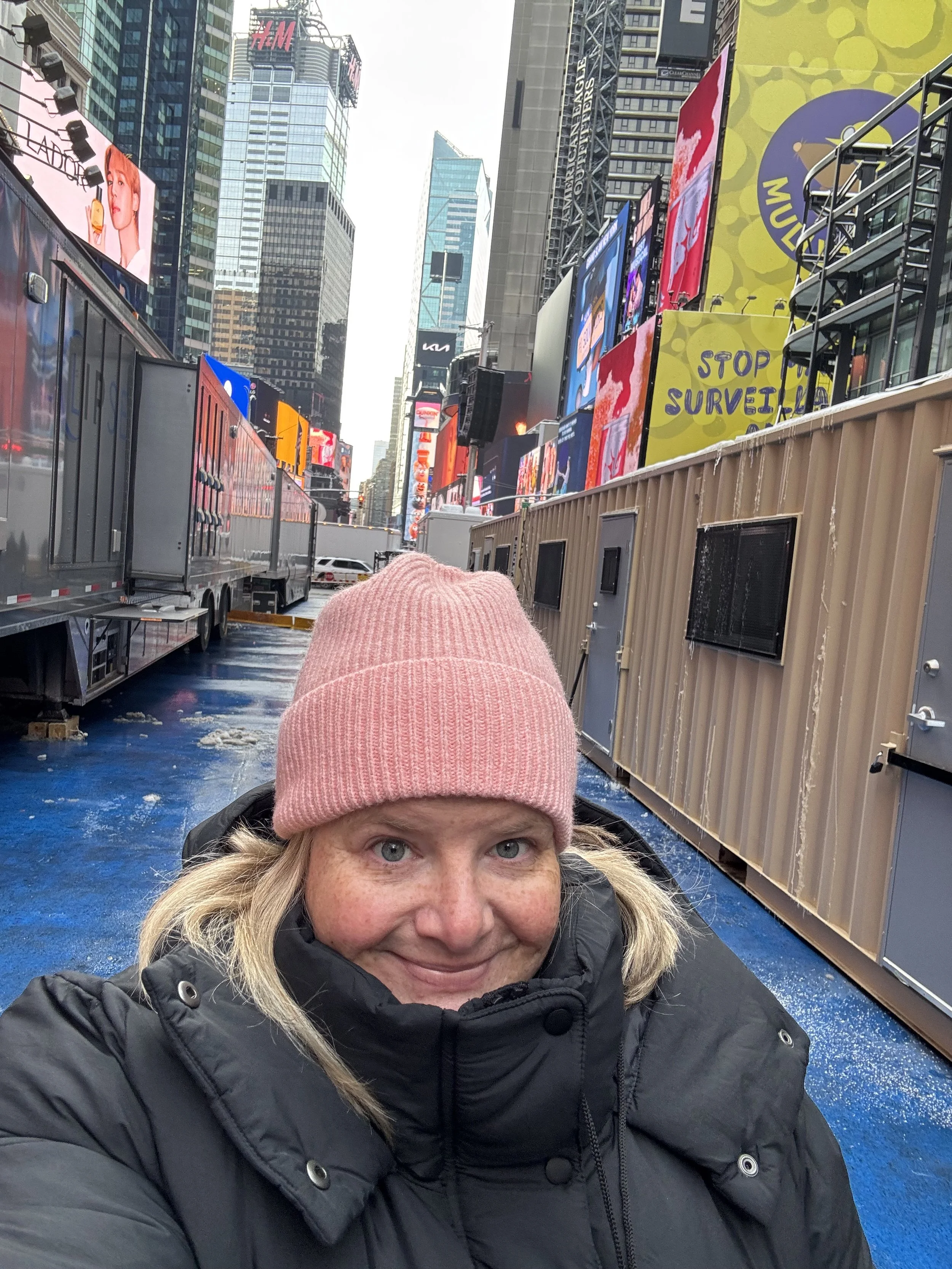 A woman in a pink knit hat and black winter coat taking a selfie in Times Square, New York City, with bright electronic billboards and tall buildings in the background.