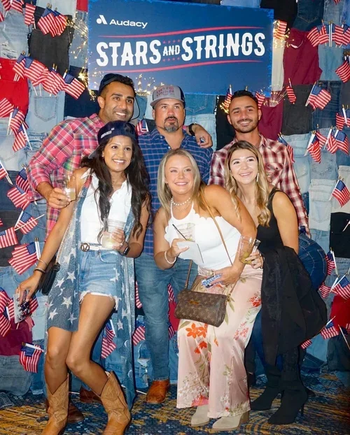 Group of six adults at a patriotic event, standing in front of a backdrop with American flags and a sign that reads 'Stars and Stripes'. They are smiling, holding drinks, and dressed in casual summer clothes.