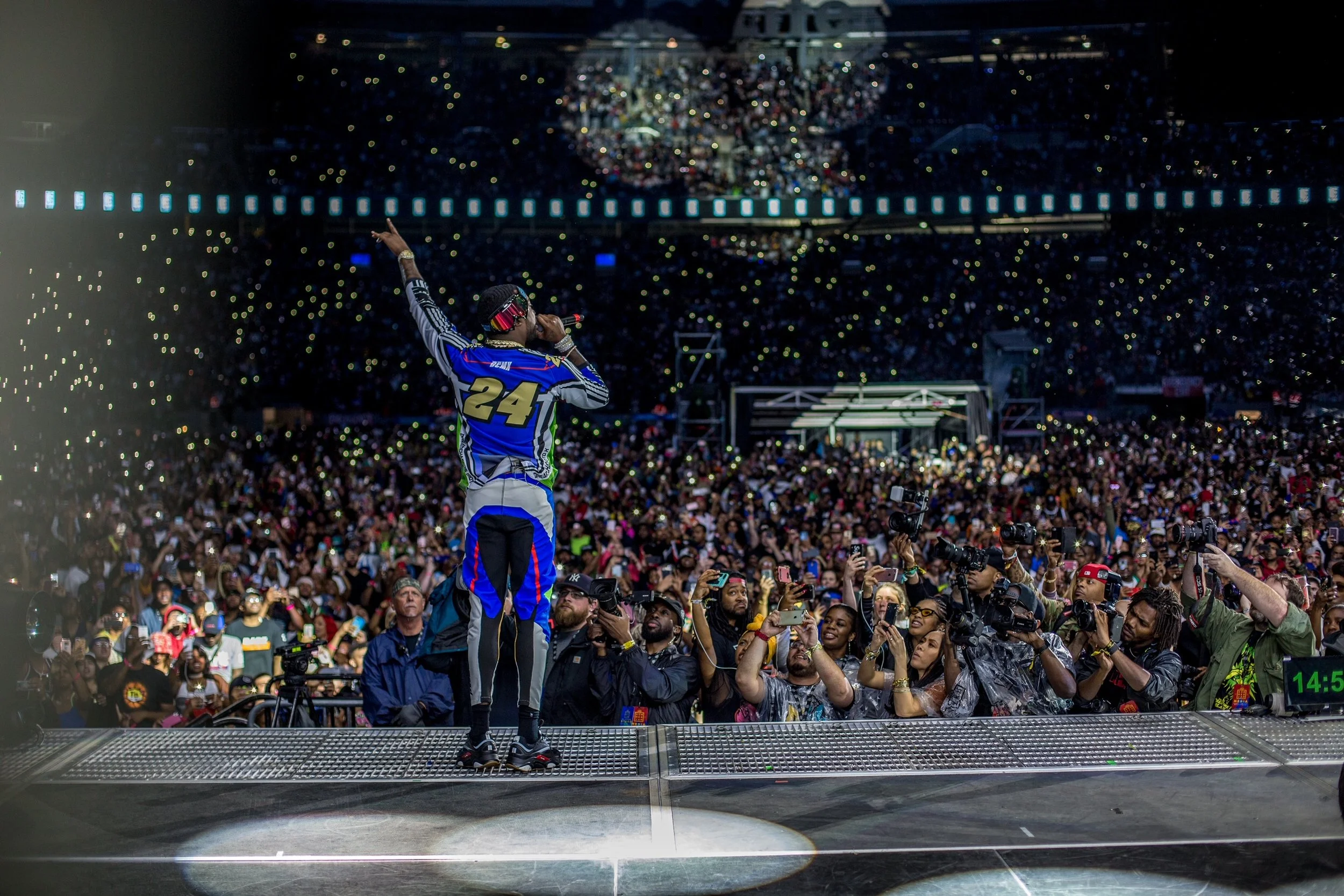 Performer on stage at a concert or music event, surrounded by a large crowd of fans and photographers, with audience lights and stage equipment visible in the background.