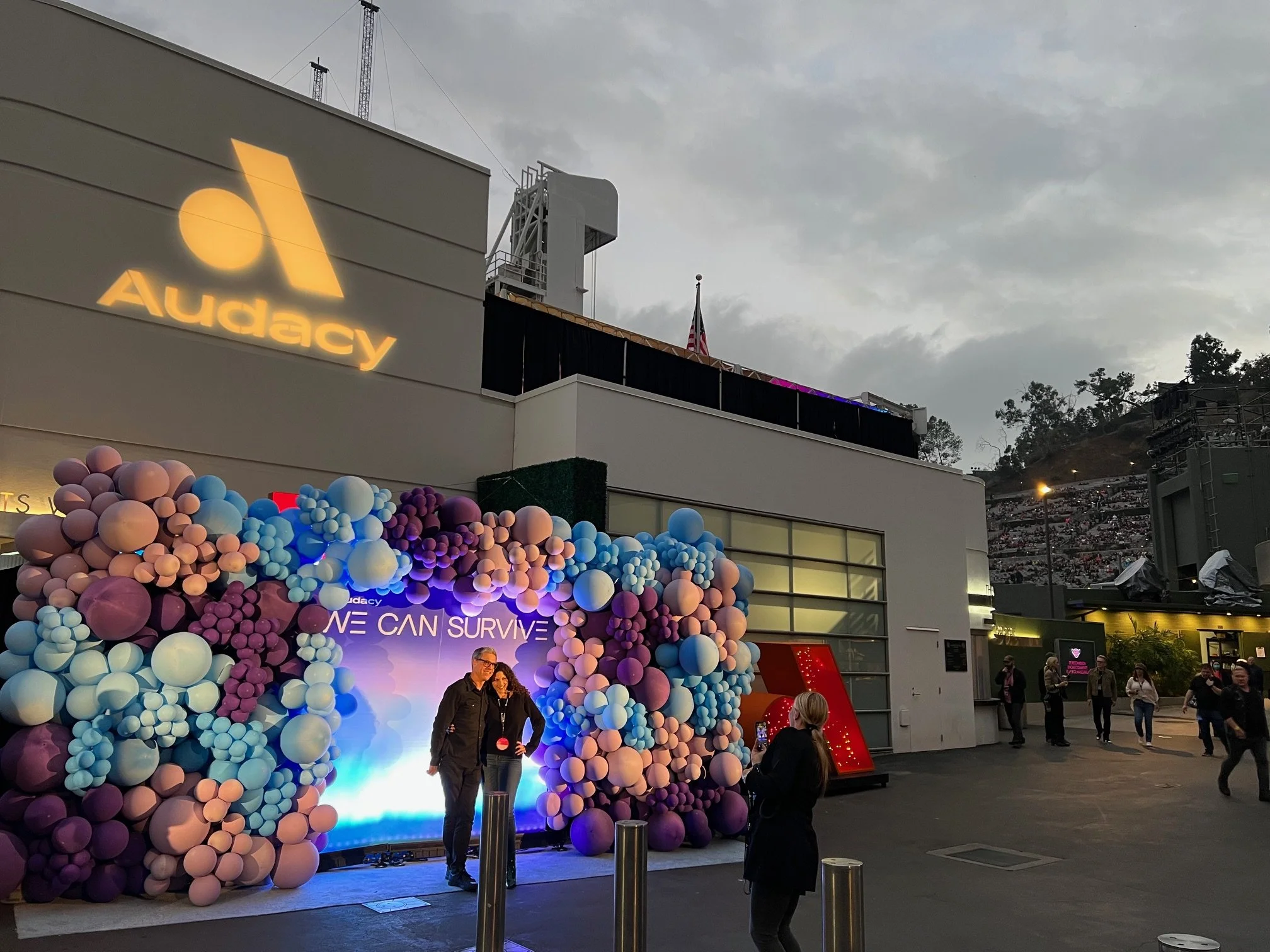 People taking photos in front of a balloon arch with a sign that reads 'WE CAN SURVIVE' at an outdoor event, with a building and an Audacy sign in the background during dusk.