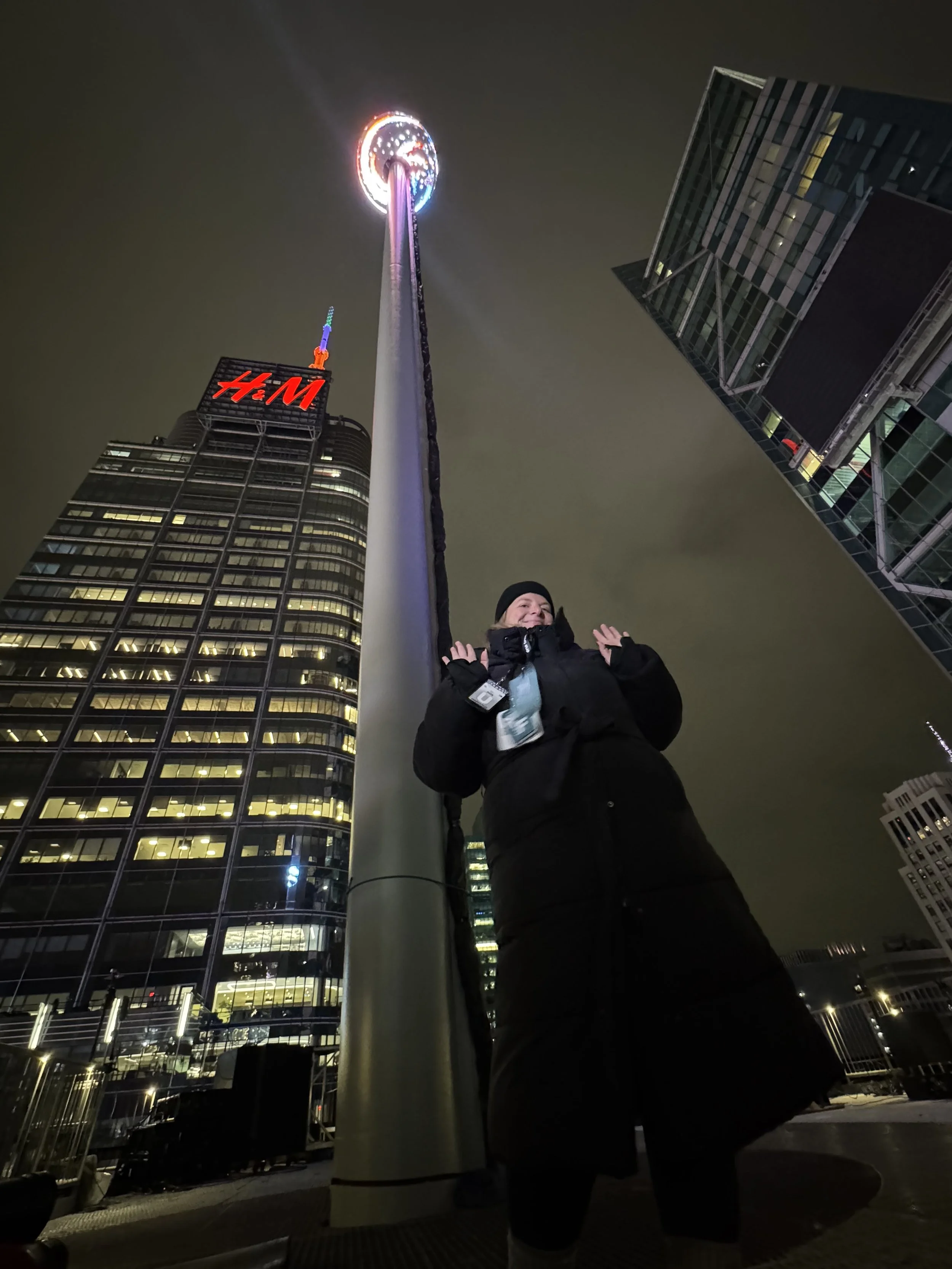 A person standing outdoors at night beneath a tall illuminated amusement park ride, with a city skyline including a building with an H&M sign in the background.