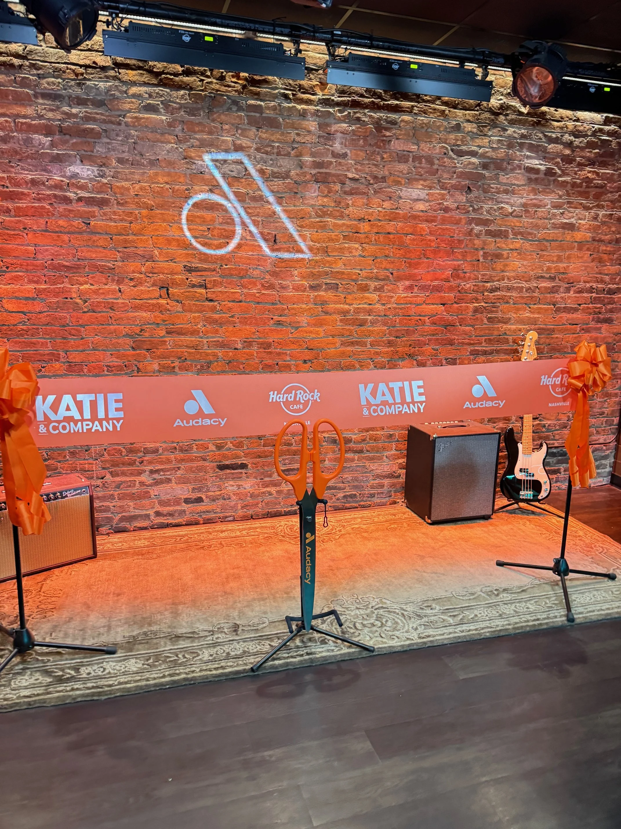 Stage with microphone stands, guitar amp, bass guitar, and event banner, set against a brick wall with a logo projected above.
