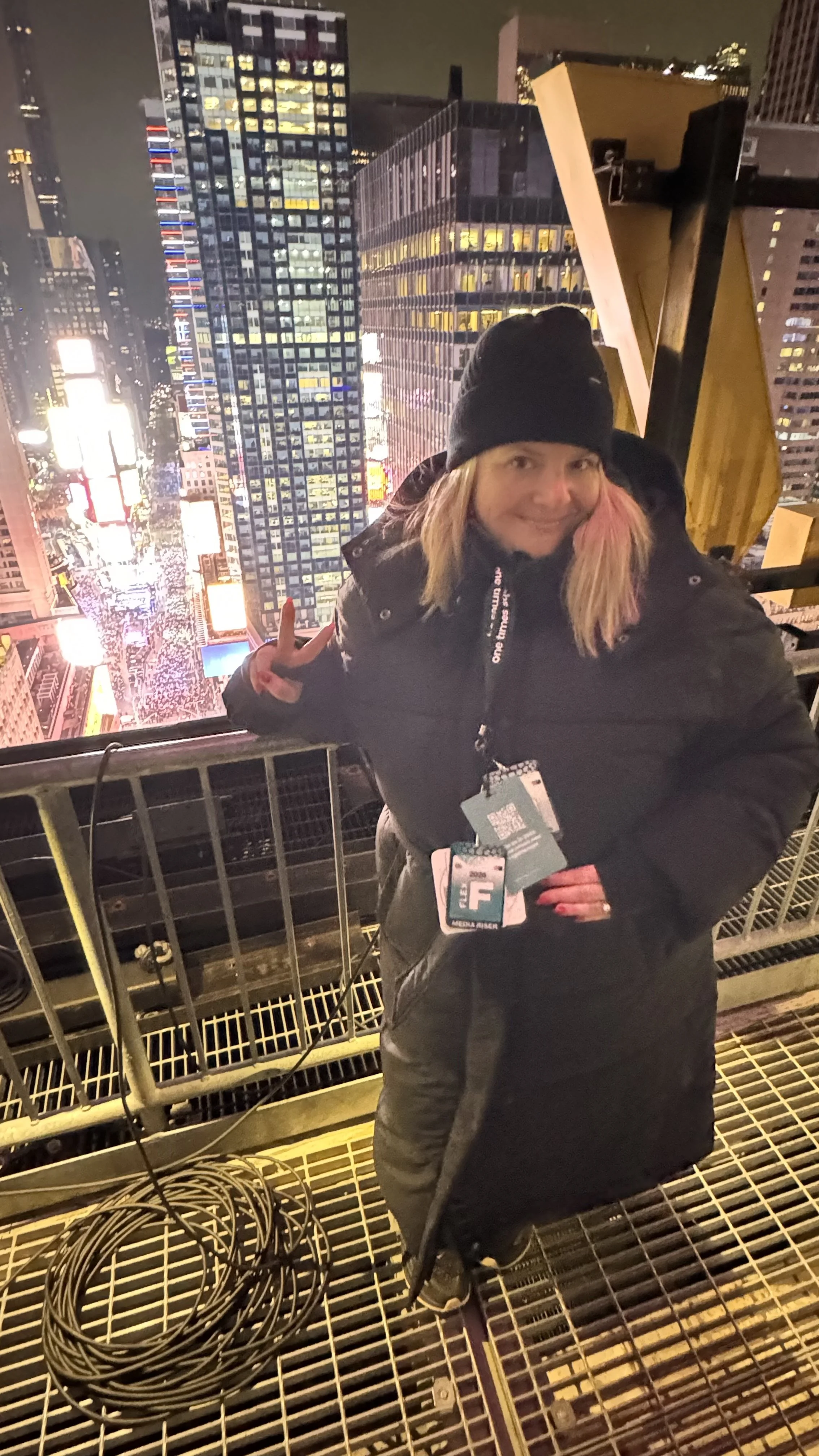 A person with blonde hair, wearing a black beanie and black winter coat, stands on a high platform in Times Square at night, flashing a peace sign with their left hand, with bright city lights and tall buildings in the background.