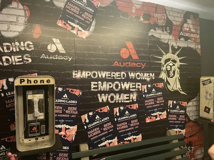 Wall with graffiti and posters promoting an event called 'Leading Ladies'. The posters feature names like Maren Morris, Bebe Rexha, Ingrid Andrews, and Jax. There is a statue of the Statue of Liberty and a bench below. A payphone is mounted on the wa