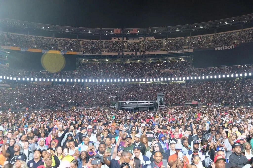 Crowd at a large stadium concert or event at night, with many people holding up phones and cameras.