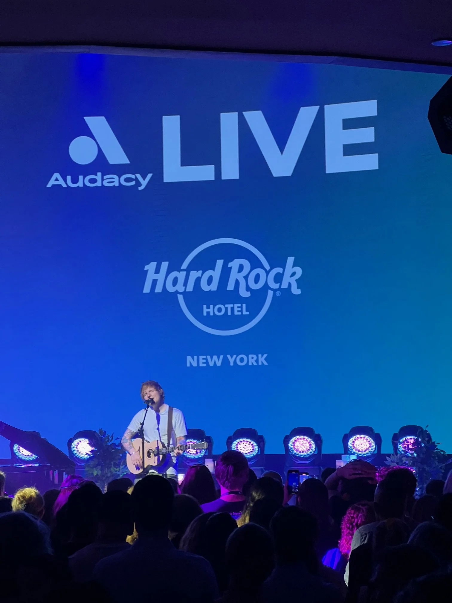 Musician performing live on stage at the Hard Rock Hotel in New York with a large screen behind displaying 'Audacy LIVE' and the Hard Rock Hotel logo.