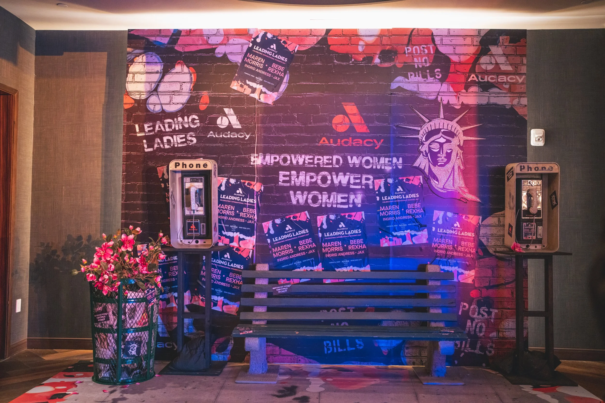A mural on a brick wall promoting women's empowerment, with posters and phrases like "Empowered Women Empower Women," and images of the Statue of Liberty. There are two payphones on either side of a black bench, a flower arrangement on the left, and 