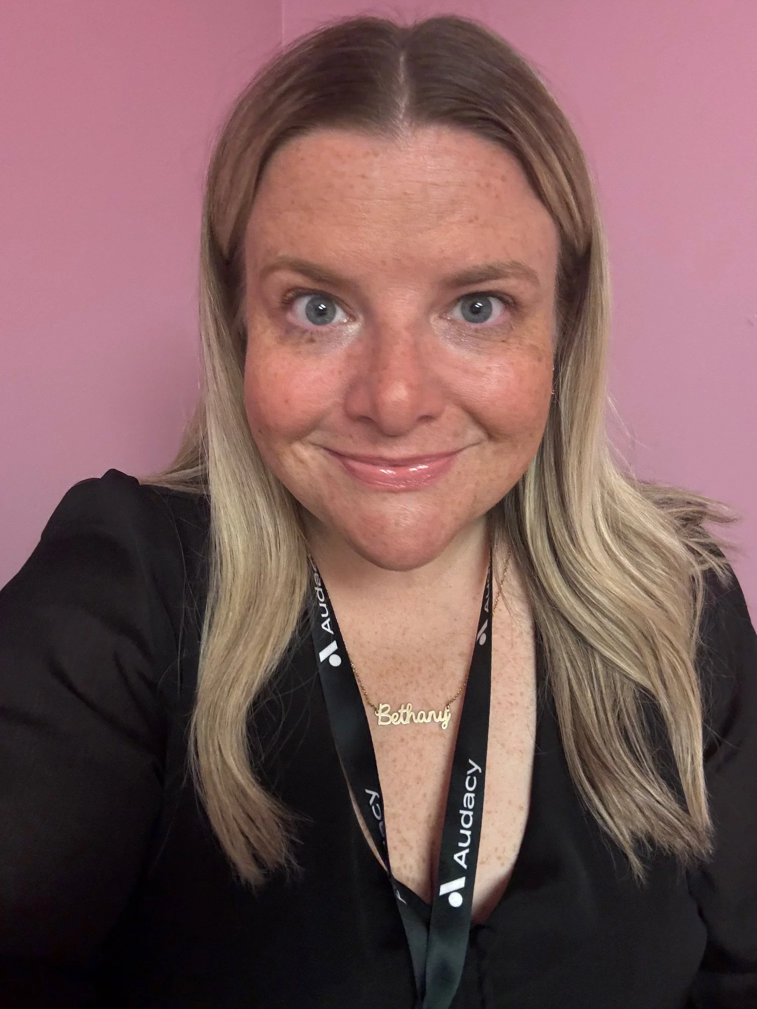 A woman with blonde hair and freckles is smiling at the camera, wearing a black top, a gold necklace with the name 'Bethany', and a black lanyard with the Audacy logo, in front of a pink background.