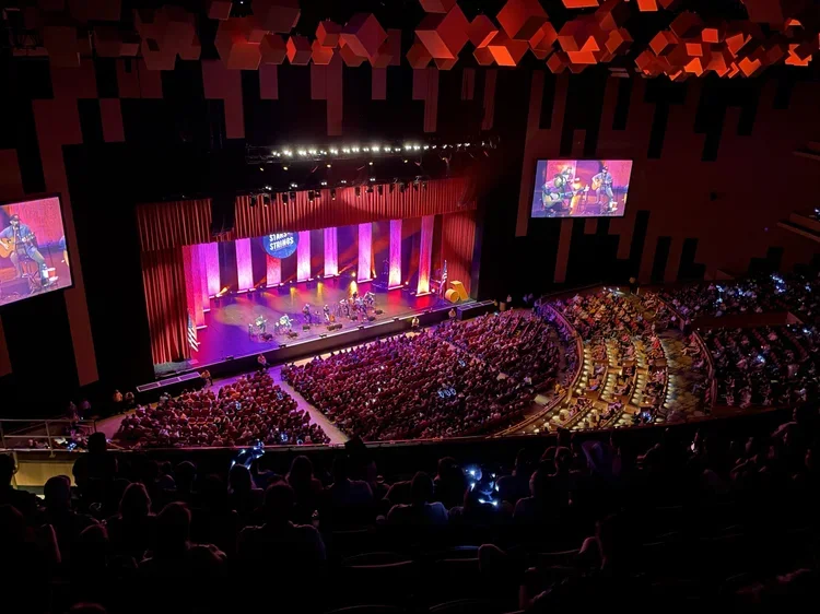 A large auditorium filled with people watching a stage performance with a band. The stage has a red curtain, colorful lighting, and two large screens displaying the performers.
