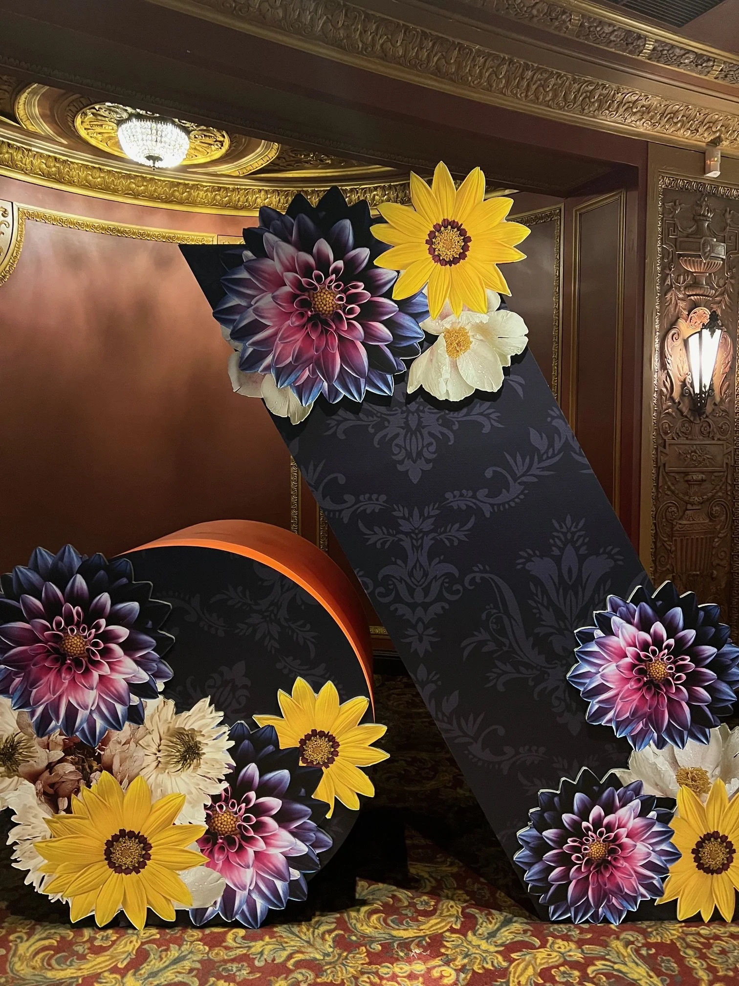 Decorative floral arrangement with large paper flowers in shades of purple, yellow, and white, placed on a dark ornamental stand in an ornate, gold-accented room.