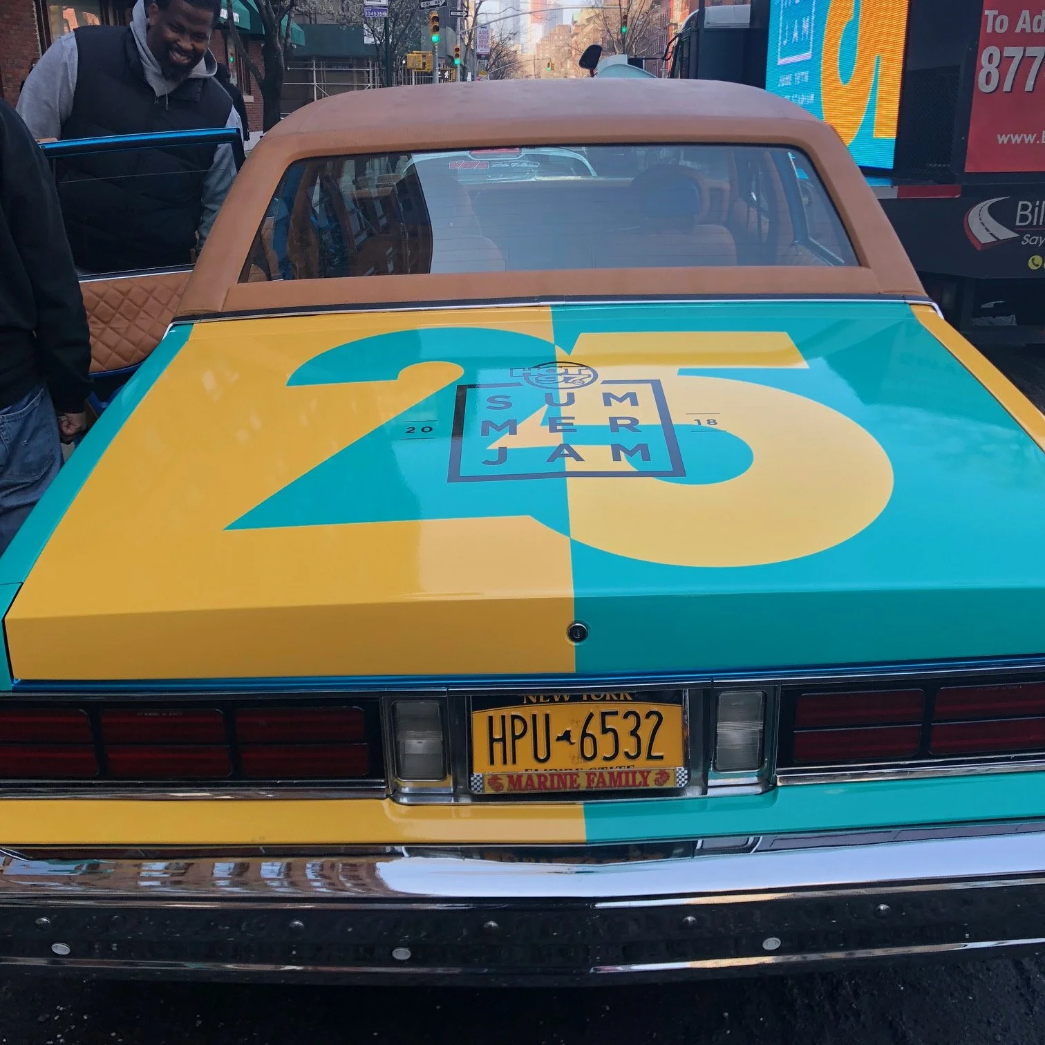 Old classic car with colorful Summer Jam 25 logo painted on the trunk, featuring large yellow and teal graphics and a New York license plate.