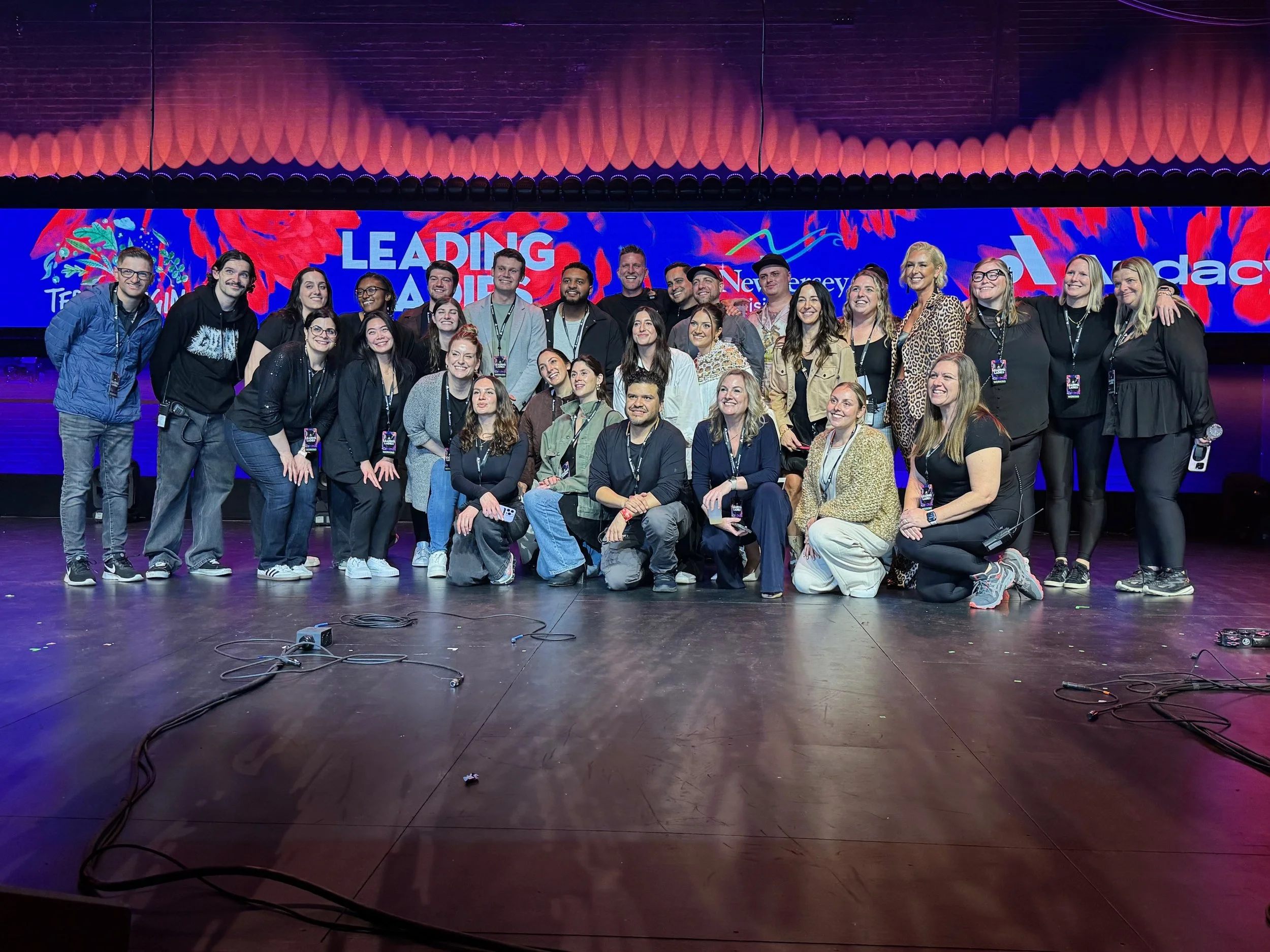 Group of diverse people on stage at an event, with a colorful background displaying the words 'Leading Audio' and logos.