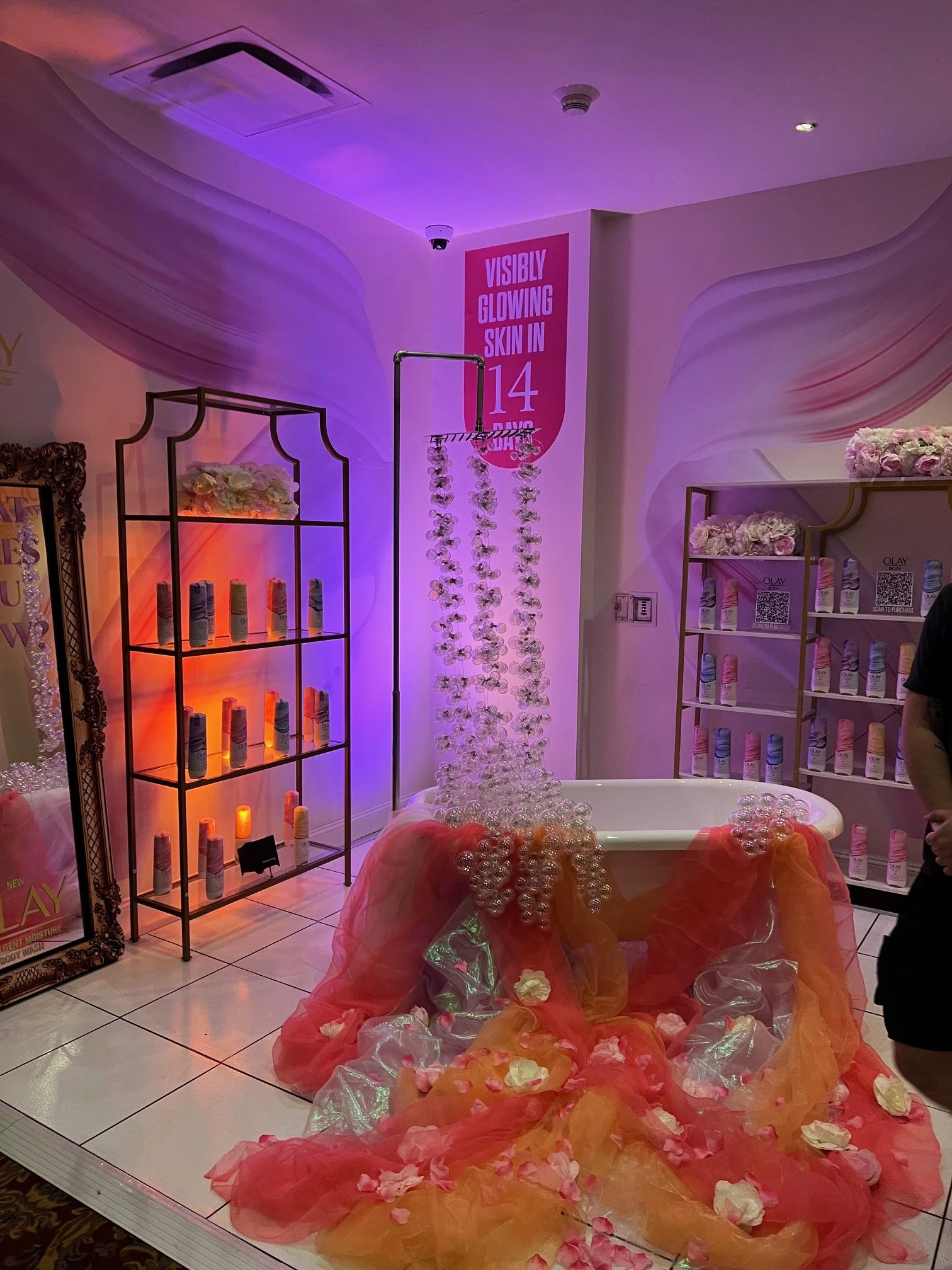 Display of skincare products at a promotional event with a luxurious bathtub surrounded by pink and orange tulle and flower petals. There is a bubble-shooting setup with hanging bubbles, banners highlighting skin benefits, and shelves of skincare bot