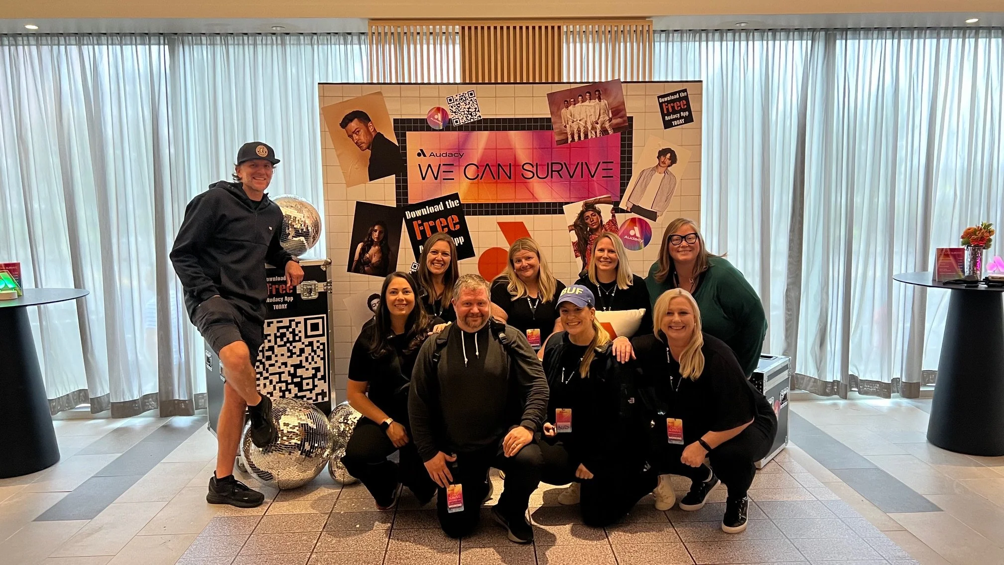 Group of nine people, including men and women, smiling and posing in front of a promotional display for Audacy's 'We Can Survive' event, with some wearing event passes and group members kneeling and standing inside a lobby area with curtains and tabl