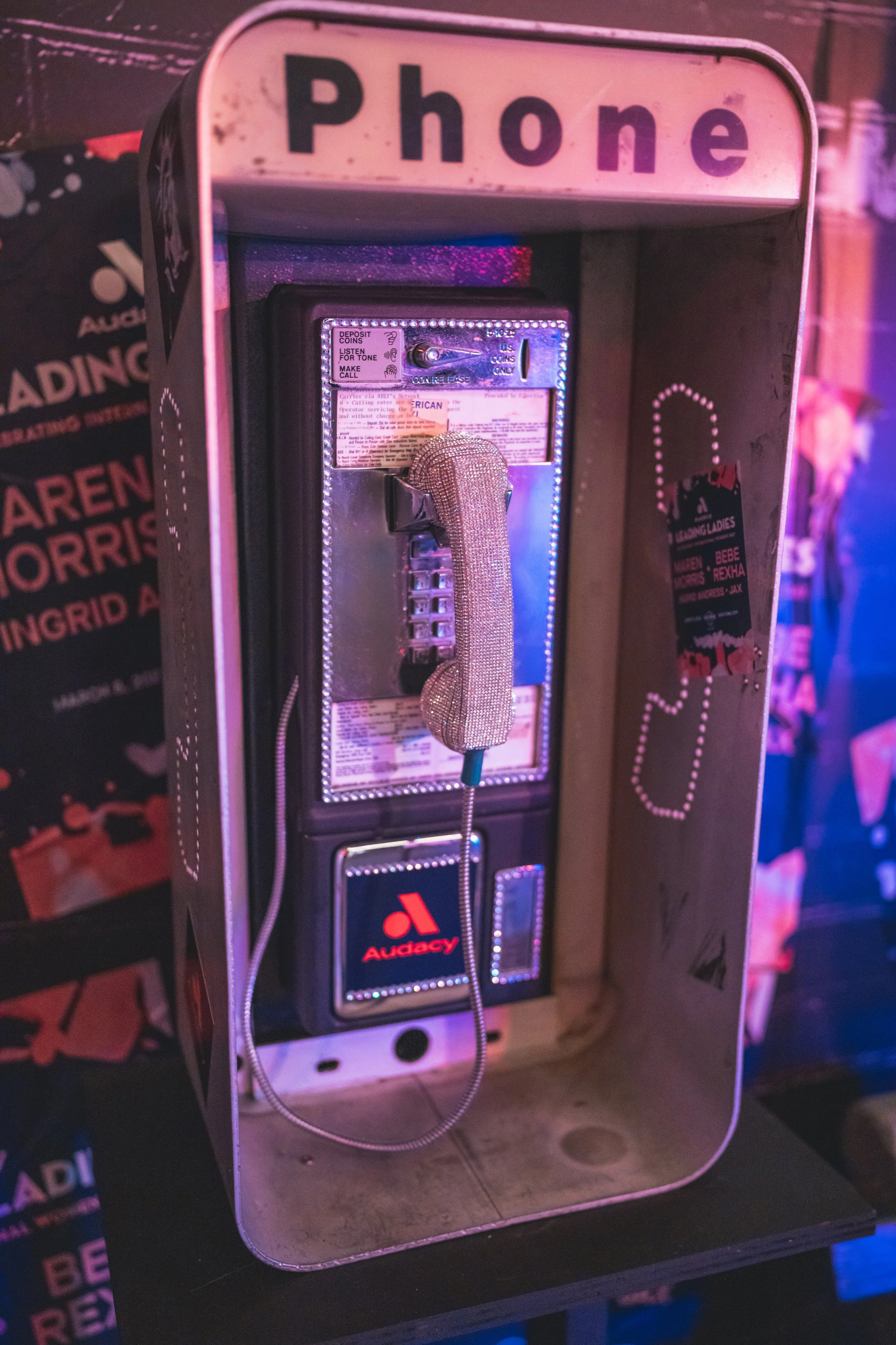 A vintage payphone inside a pink metal phone booth with the word 'Phone' on top, decorated with neon lights and a glittery, diamond-encrusted handset.