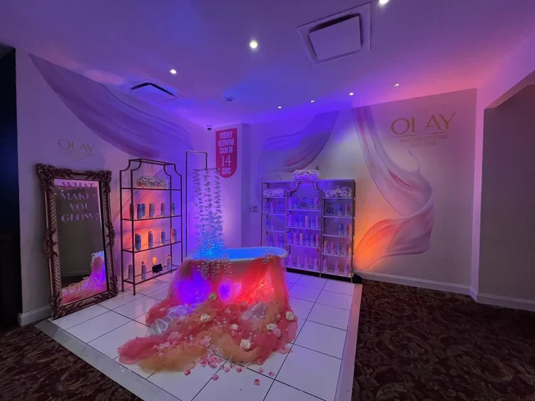 A spa or beauty product display area with pink and purple lighting, featuring a large mirror, shelves with products, a bathtub, and a backdrop with the Olay logo.