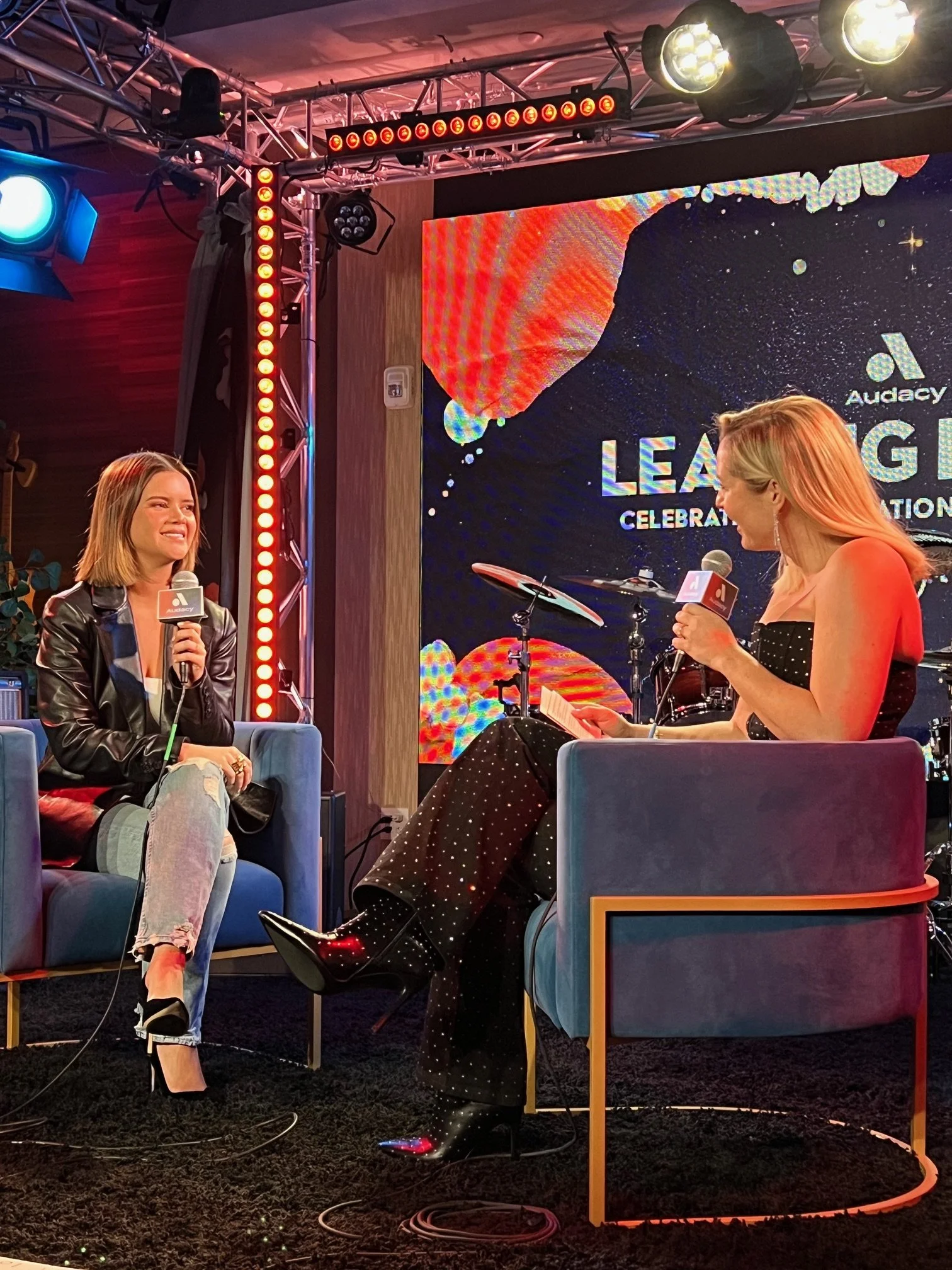 Two women are sitting on stage in conversation, both holding microphones. One woman has short brown hair and is dressed in a black leather jacket and ripped jeans. The other woman has long blonde hair, is dressed in a Freda dress, and is looking at h
