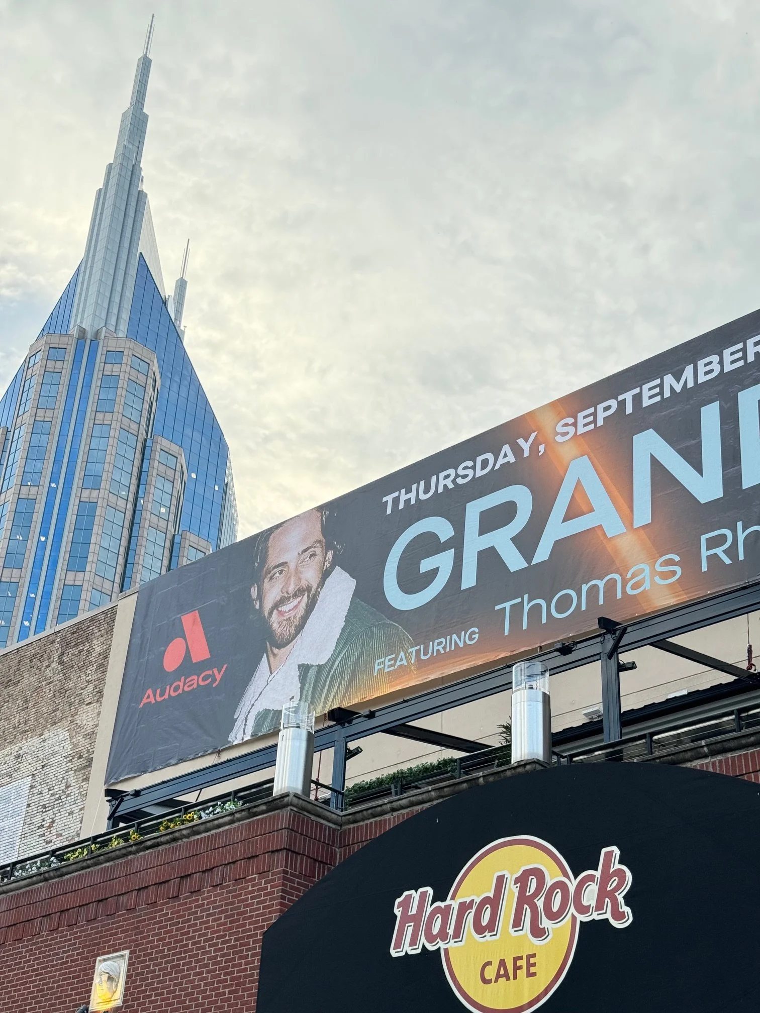 Urban scene with a large billboard advertising an event featuring Thomas Rh, and a Hard Rock Cafe sign in the foreground, with a modern blue skyscraper in the background.