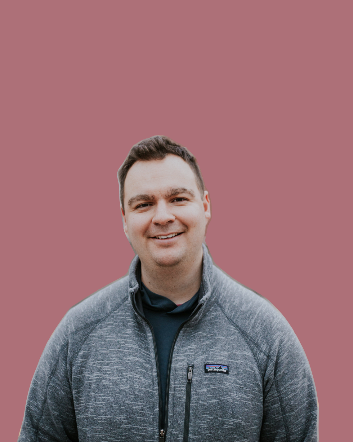 A smiling man with short dark hair wearing a gray Patagonia jacket and black shirt against a solid pink background.