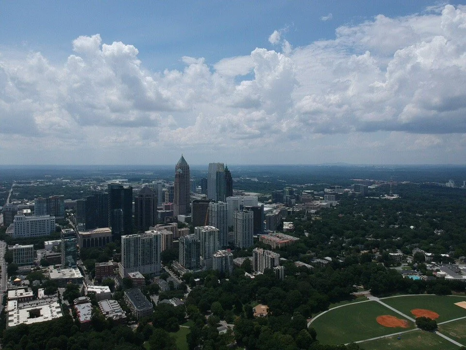 Aerial view of a city skyline with tall skyscrapers and a park with baseball fields in the foreground, under a partly cloudy sky.
