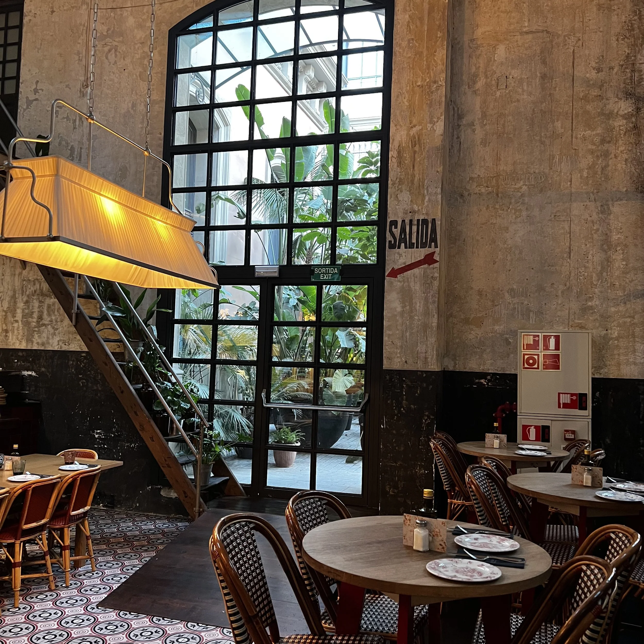 Interior of a restaurant with wooden tables and woven chairs, a large window with plants outside, a staircase nearby, and fire safety equipment on the wall.