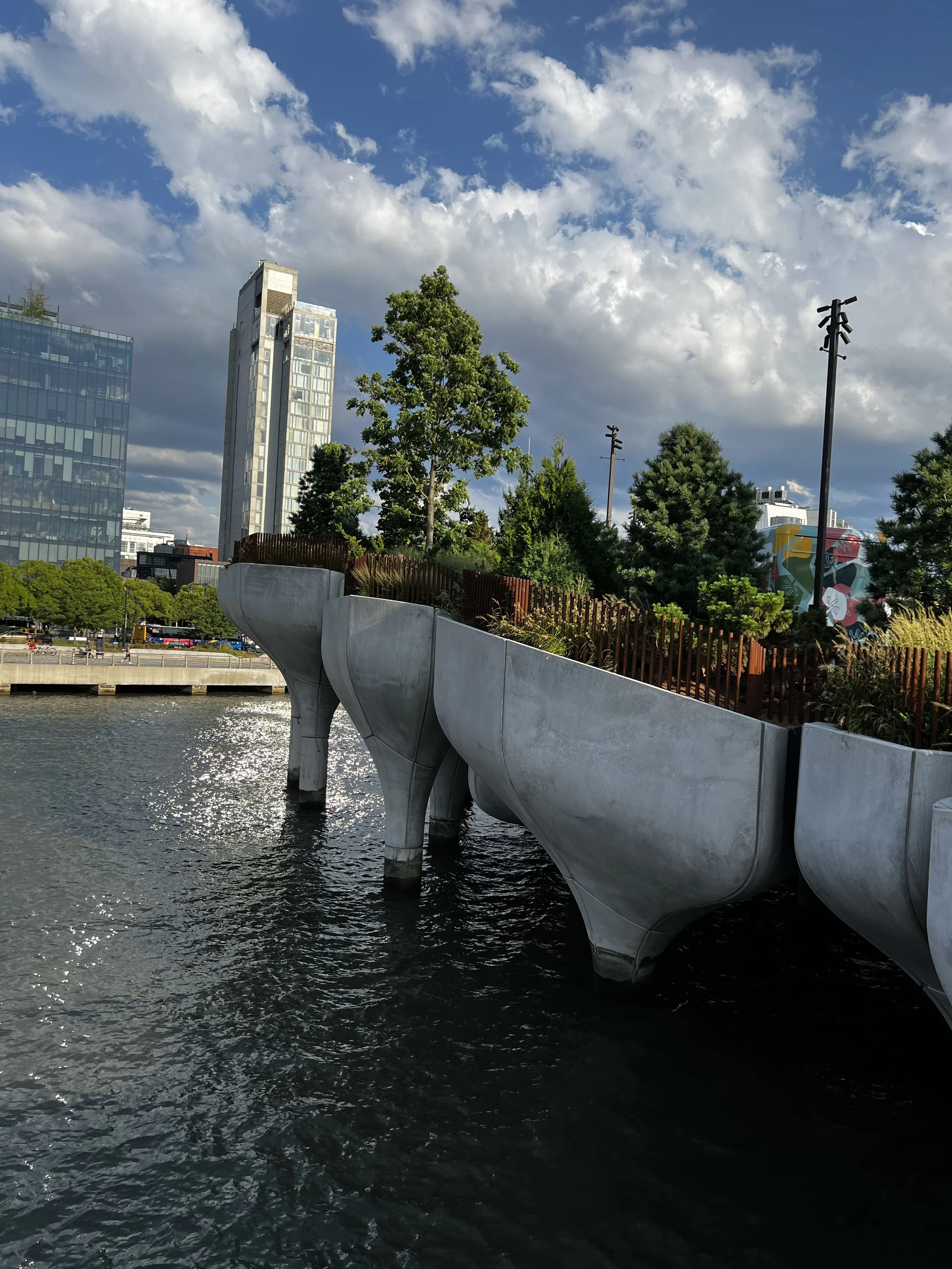Modern outdoor water feature with concrete, high-standing planters holding trees and shrubs, along a river or waterfront, with city buildings and a partly cloudy sky in the background.