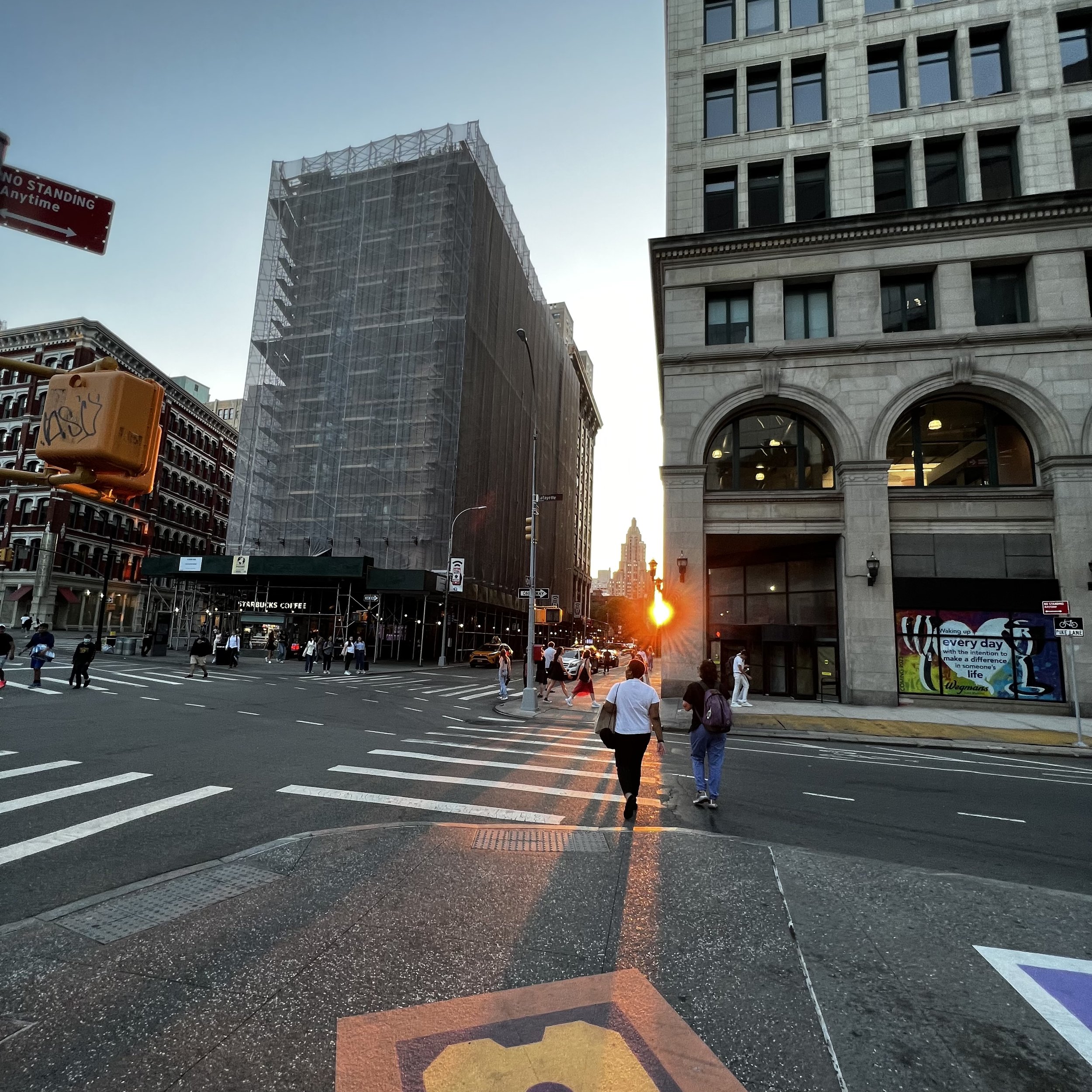 City street at sunset with pedestrians crossing. Tall buildings with windows, some under construction. Sunset sun visible in the background.