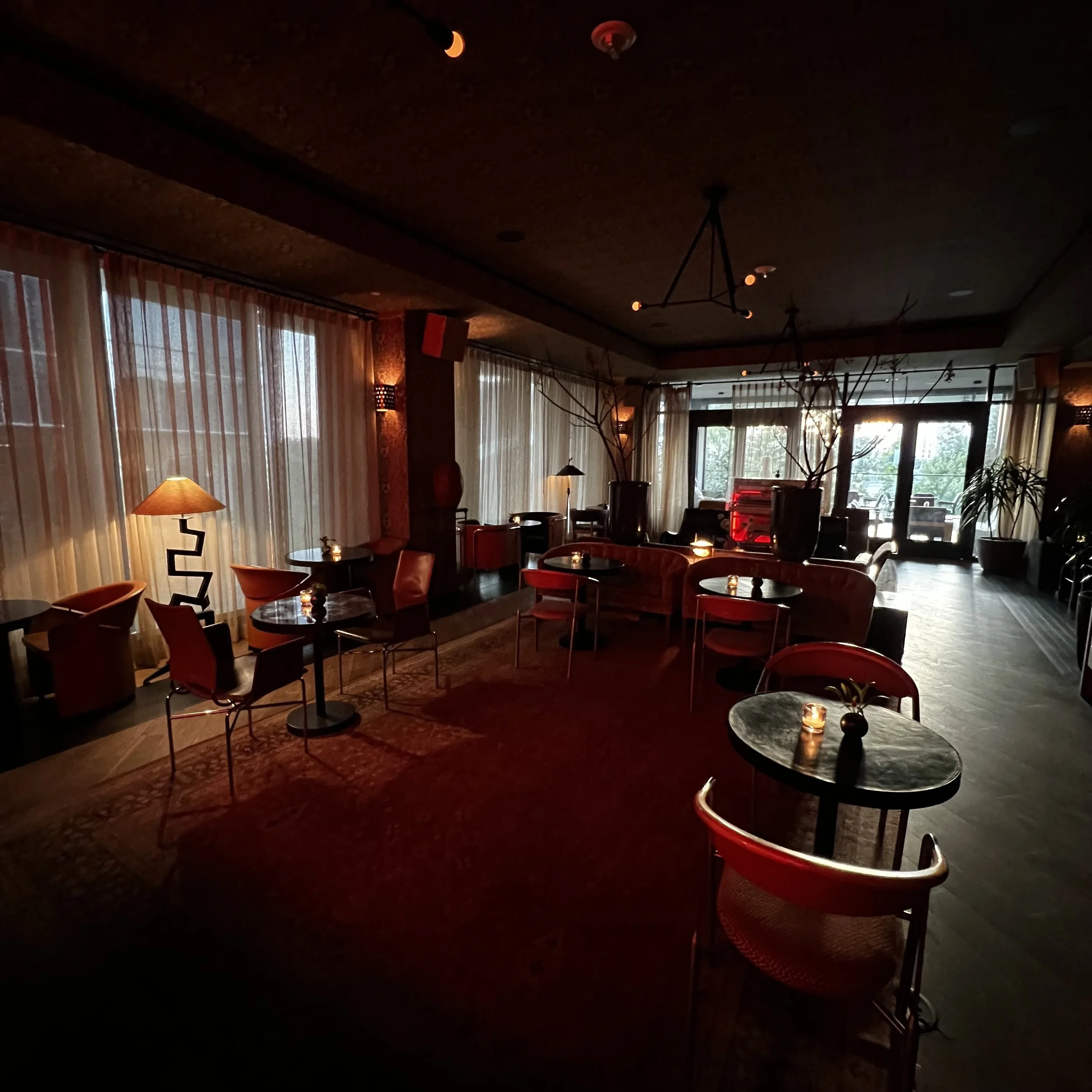 Dimly lit lounge with round tables, chairs, and large windows with curtains, decorated with plants and modern lights.