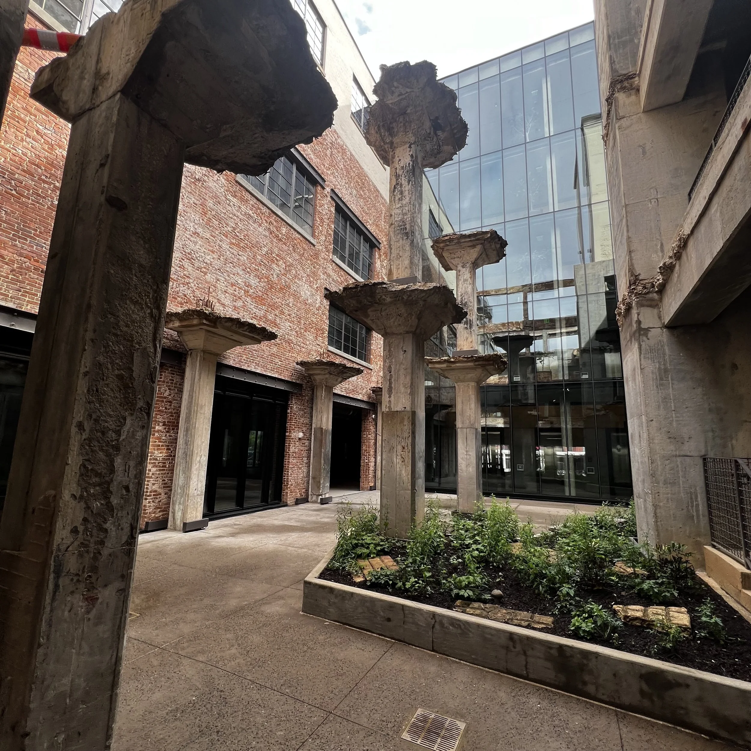 Outdoor courtyard with a garden in a rectangular wooden planter box surrounded by exposed brick and concrete buildings. There are several large, weathered concrete columns with flat, thick tops, and the background features modern glass windows.