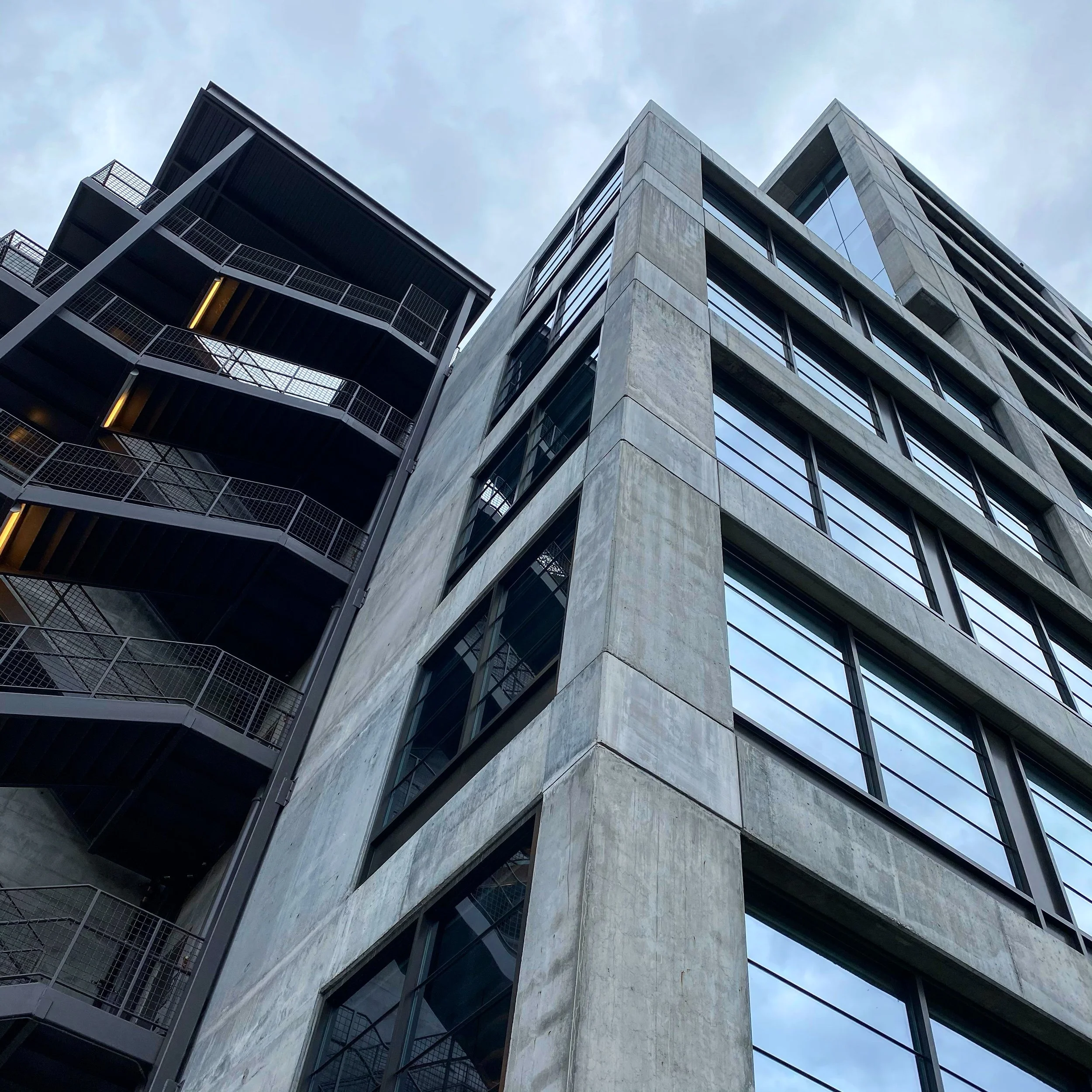 Low-angle view of modern high-rise building with concrete facade and large glass windows next to a building with black metal staircases and railings.