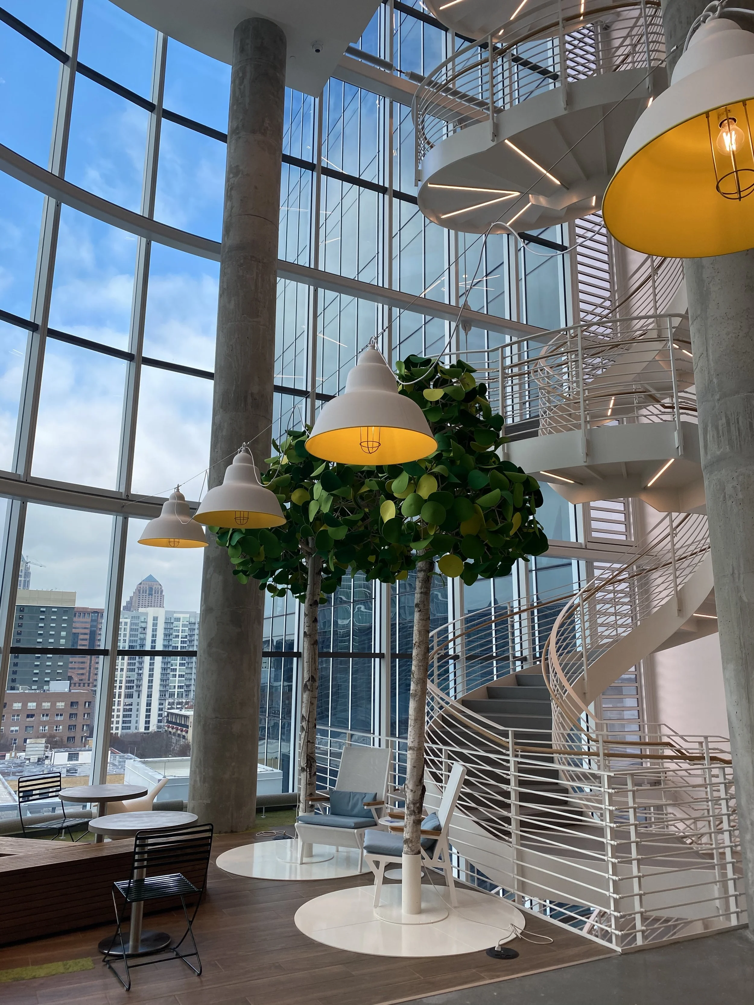 Interior view of a modern building with large glass windows, a spiral staircase, hanging yellow lamps, and a small seating area with chairs and tables.