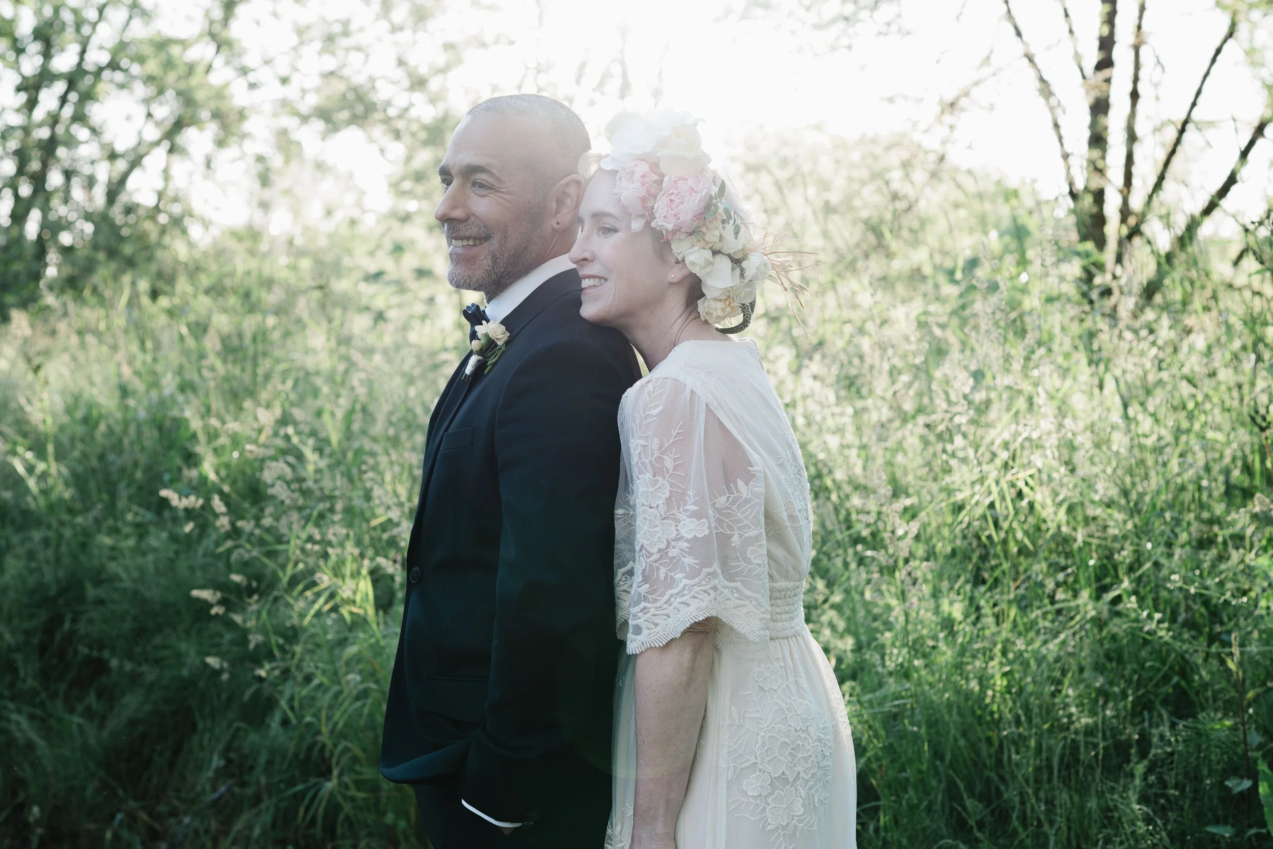 Outdoor wedding portrait of smiling bride and groom standing near sunlit meadow surrounded by tall green grass and trees.