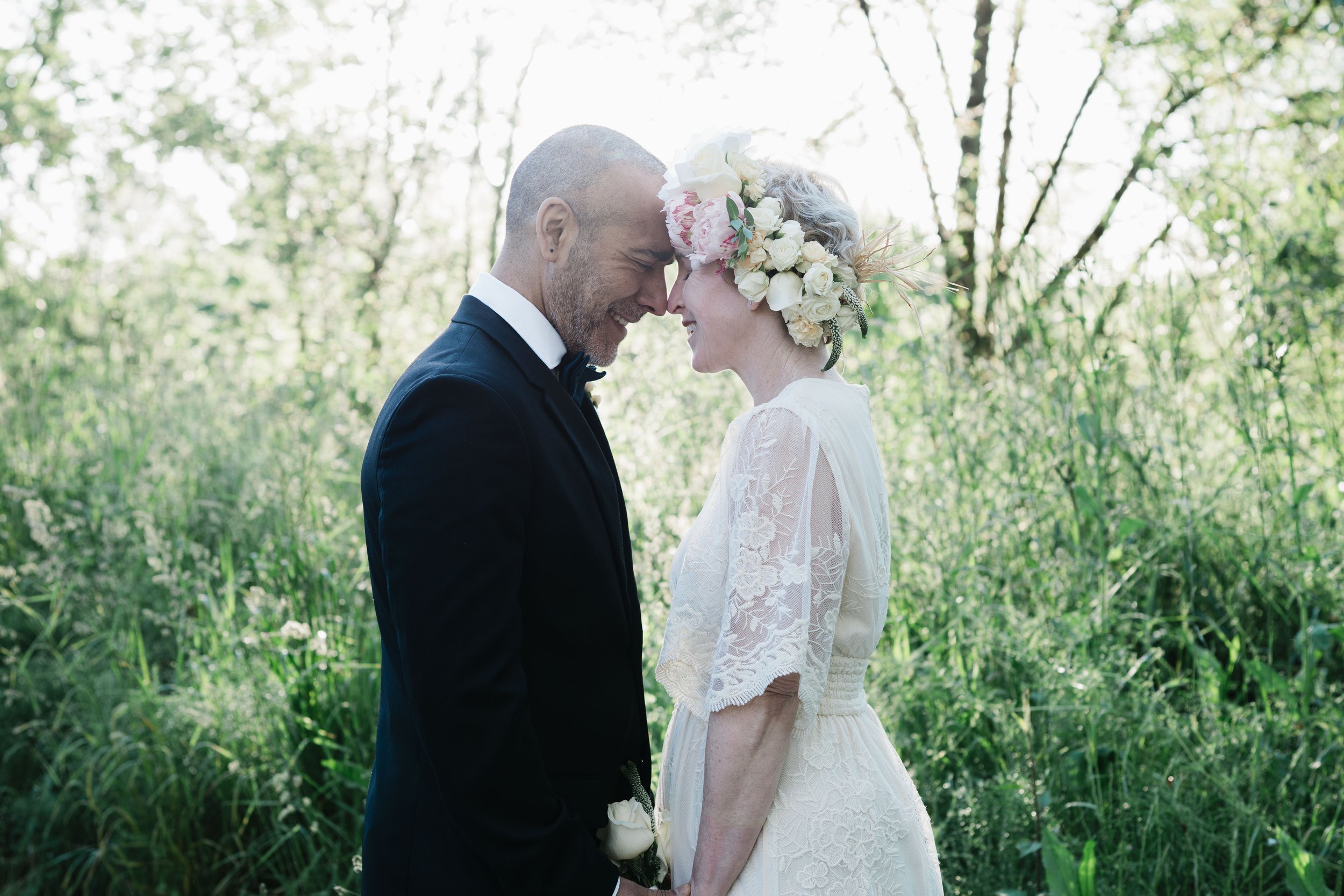 Bride and groom standing forehead to forehead in a sunlit meadow surrounded by tall grass and trees during an outdoor wedding portrait session