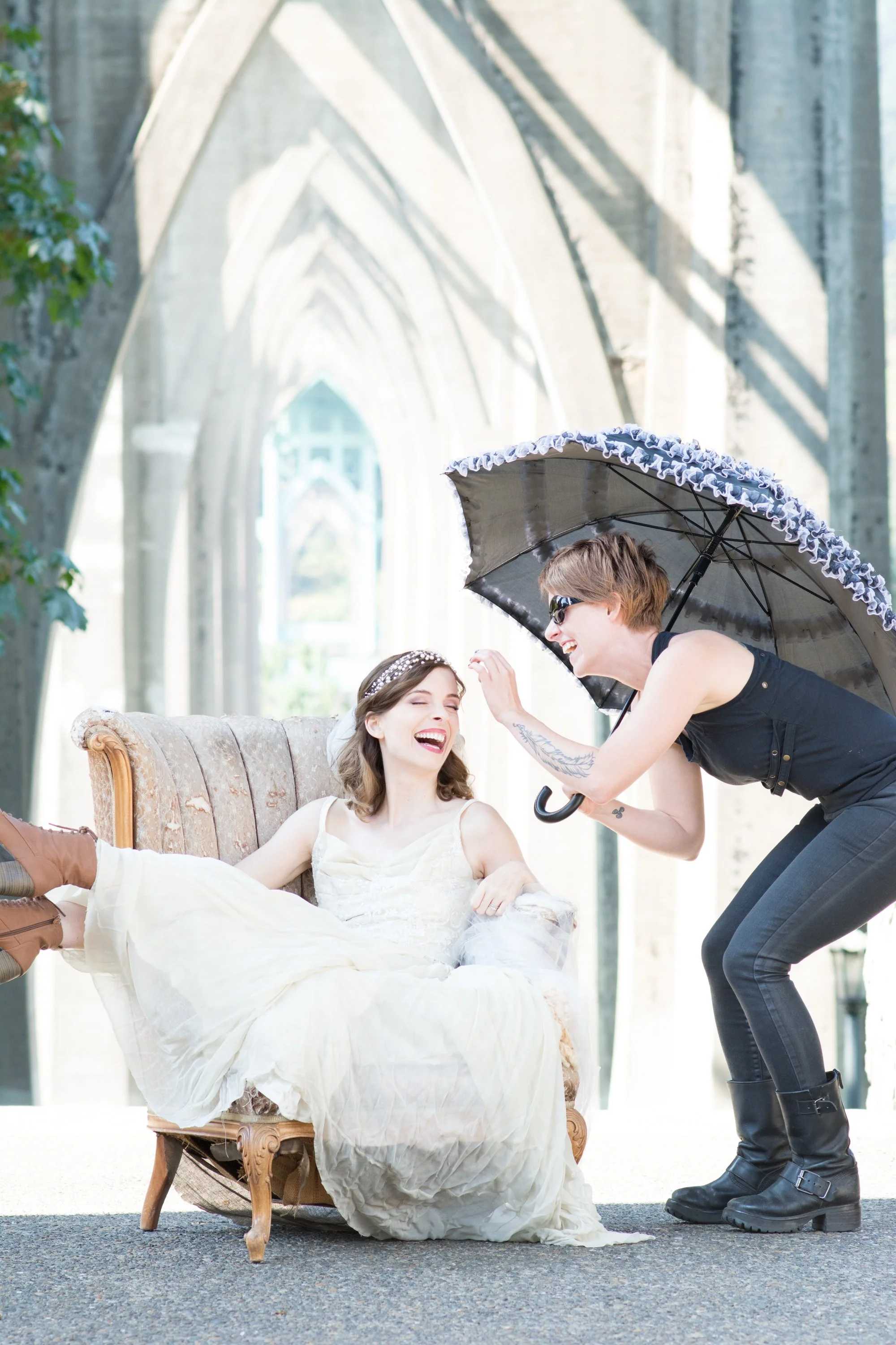 Behind the scenes image of bride sitting on vintage chair laughing with makeup artist.  Beautiful sun filled day under St. Johns Bridge in Portland Oregon