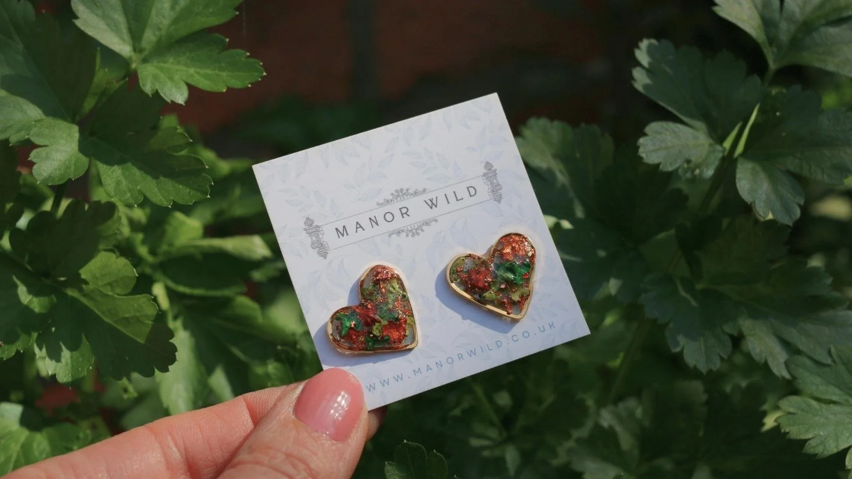 A hand holding a card with gold heart-shaped earrings on it, labeled 'Manor Wild', against a backdrop of green leafy plants.