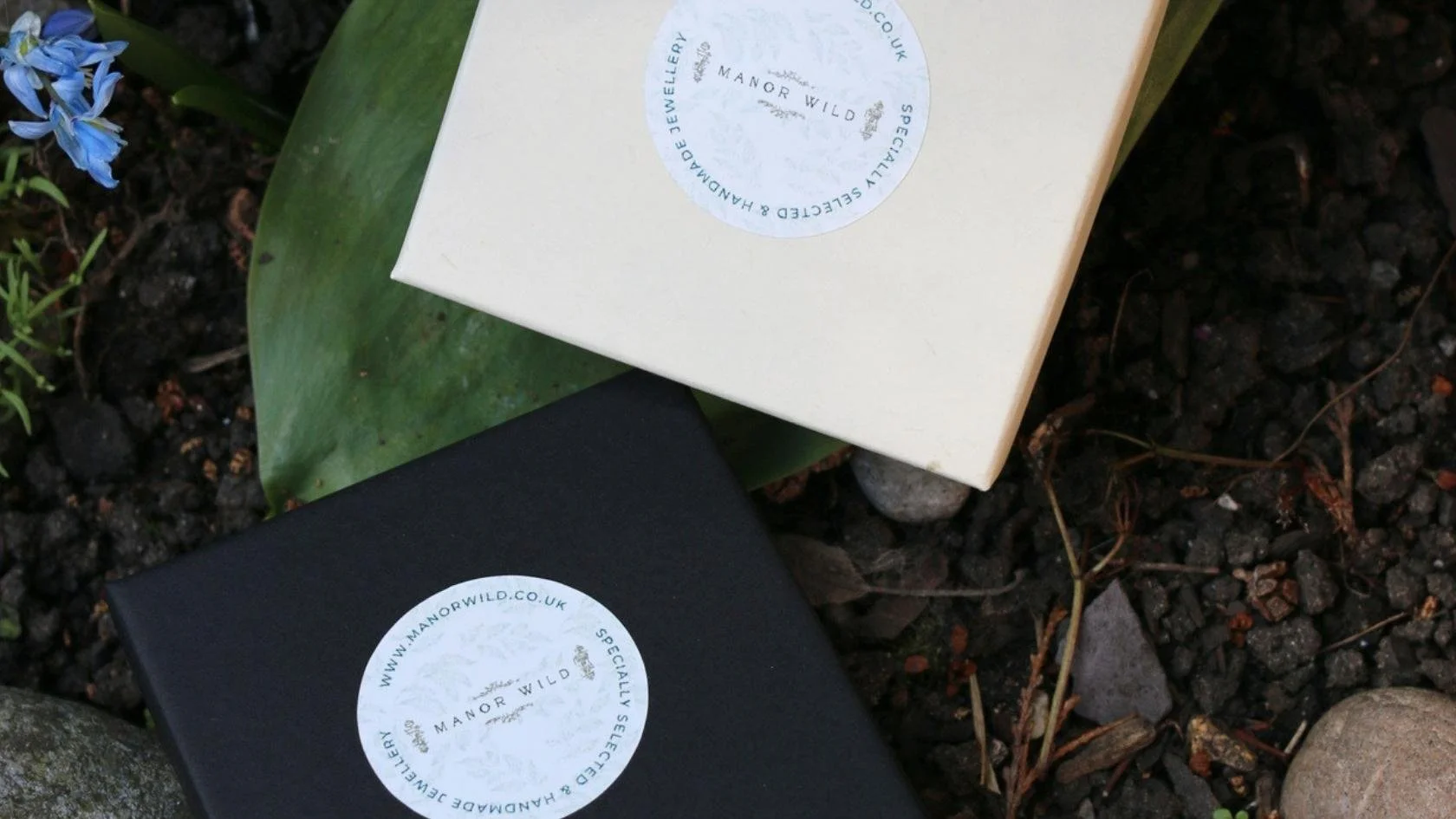Two boxes, one black and one cream, with round labels that read 'Manor Wild' and 'Specially Selected Handcrafted Jewellery,' resting on soil ground with small rocks, dried twigs, and a green leaf, and a small blue flower nearby.