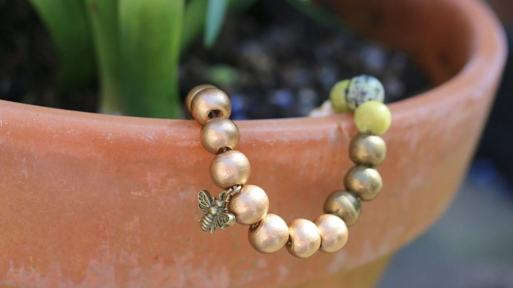 Gold-colored beaded bracelet with a small bee charm, draped over the edge of a terracotta flowerpot containing a green plant.
