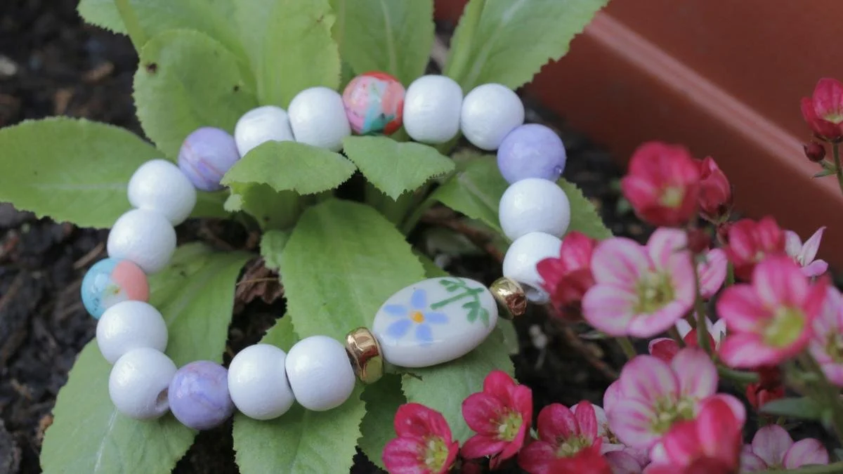 Colorful beaded bracelet with white, purple, and pastel beads, resting on green leaves next to pink flowers in garden soil.