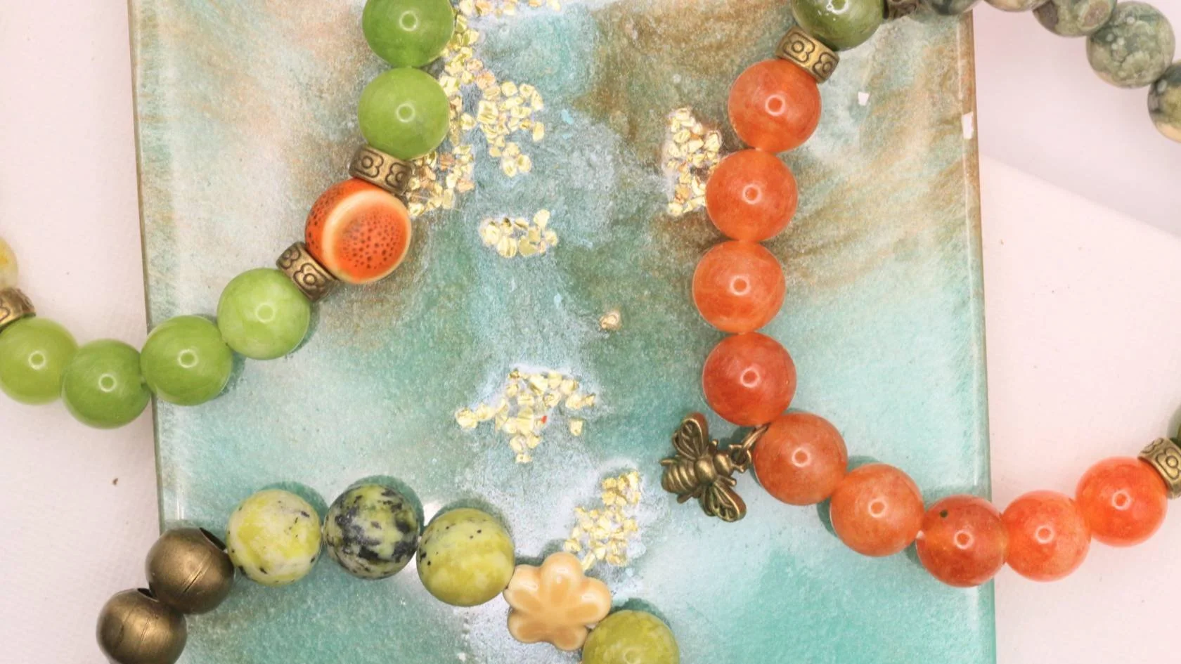 Colorful beaded necklaces arranged on a textured surface, featuring green, orange, and multicolored beads, some with decorative patterns and metallic accents.