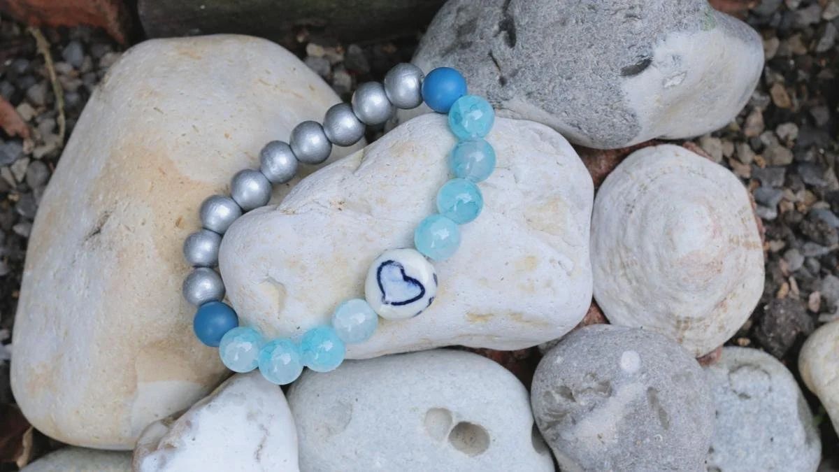 A beaded bracelet with blue, silver, and white beads, and a white bead with a hand-drawn blue heart, resting on white and gray rocks.