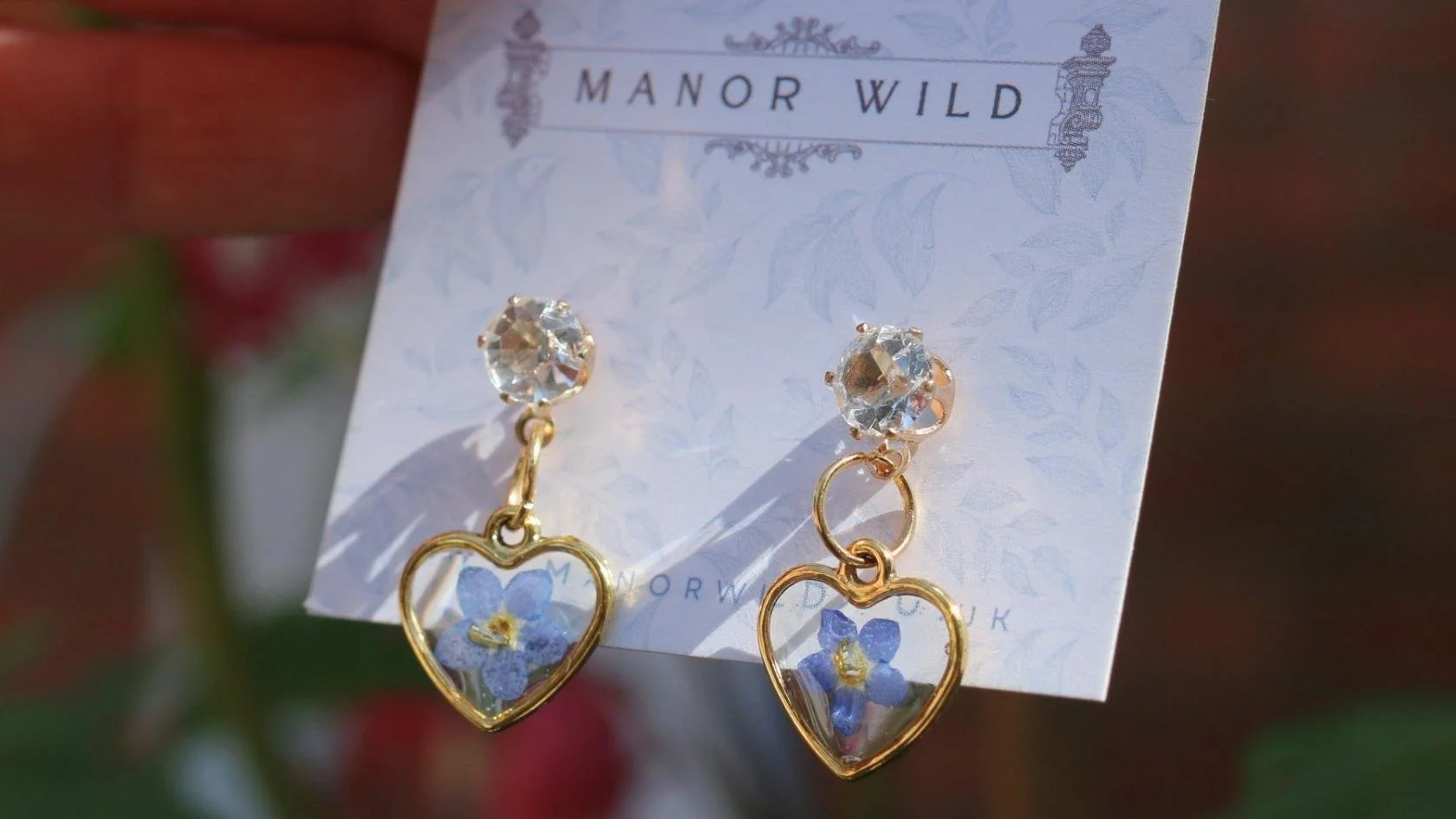 Pair of gold earrings with heart-shaped pendants containing blue flowers and clear gemstones, displayed on a card labeled 'Manor Wild'.