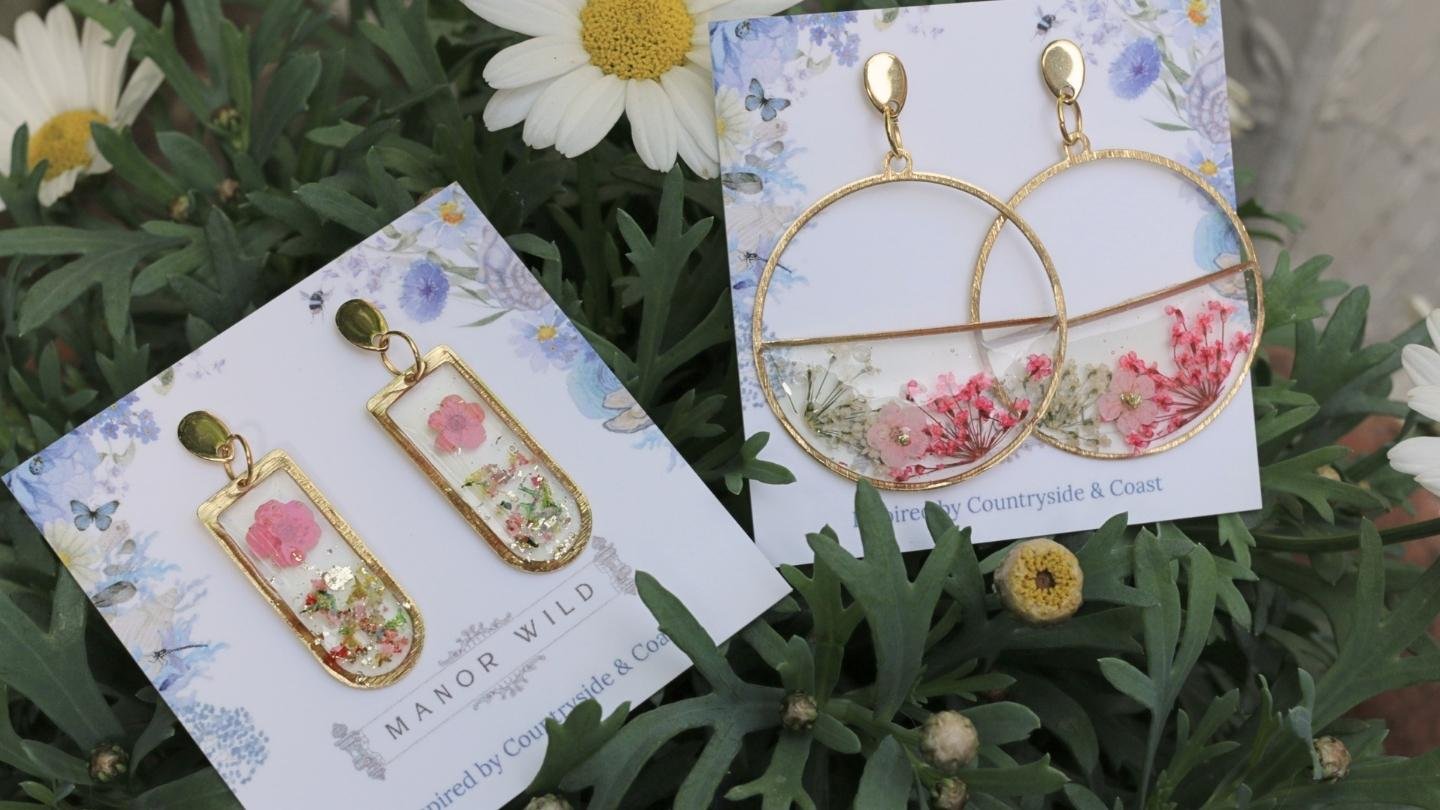 Two pairs of earrings displayed on floral patterned cards, placed on green foliage. The first pair are rectangular with gold frames and pink flowers inside. The second pair are large circular hoops with gold rims, also containing pink and other small